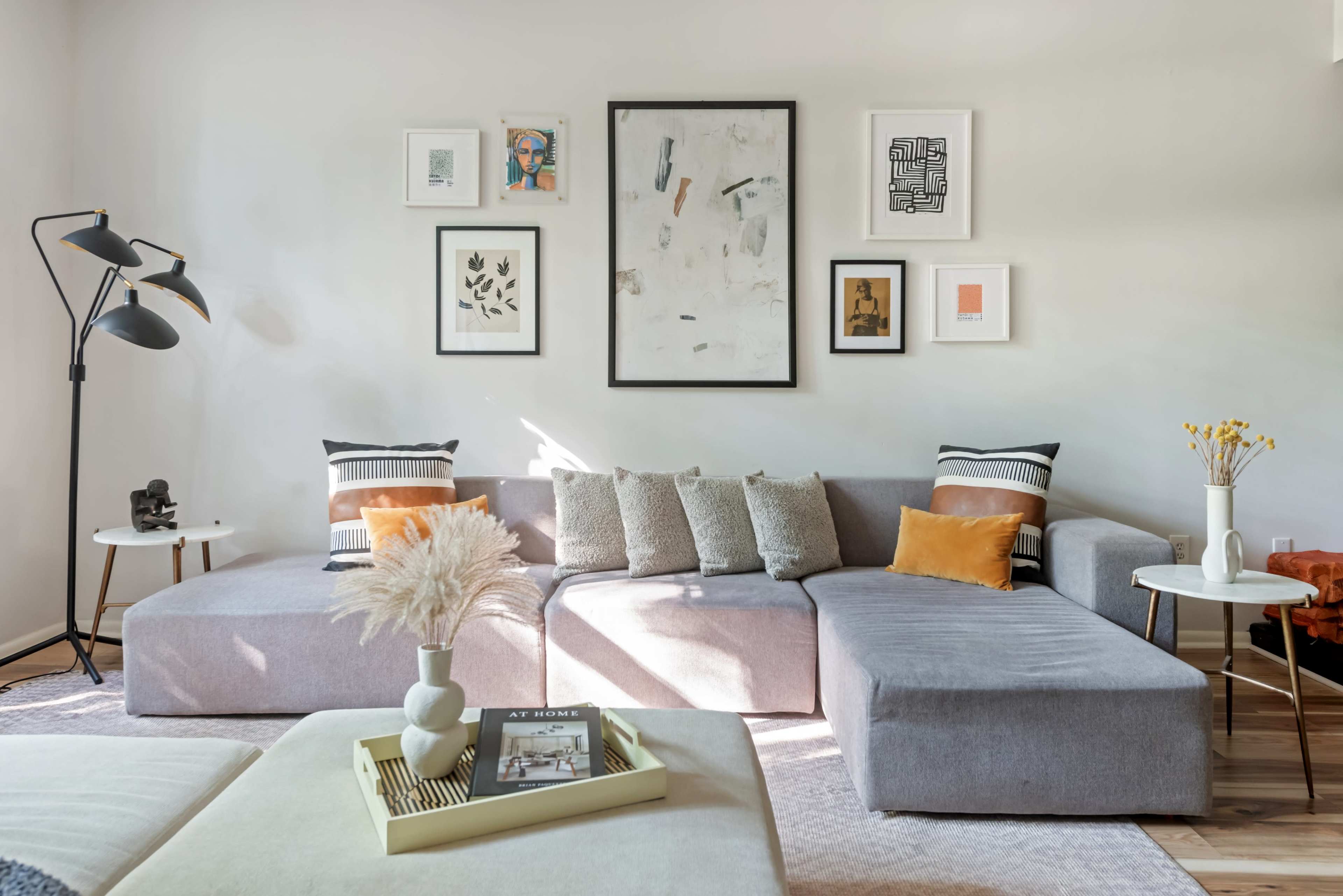 A light-filled living room features a gray sectional sofa adorned with decorative pillows, framed artwork on the walls, and a floor lamp in the corner.