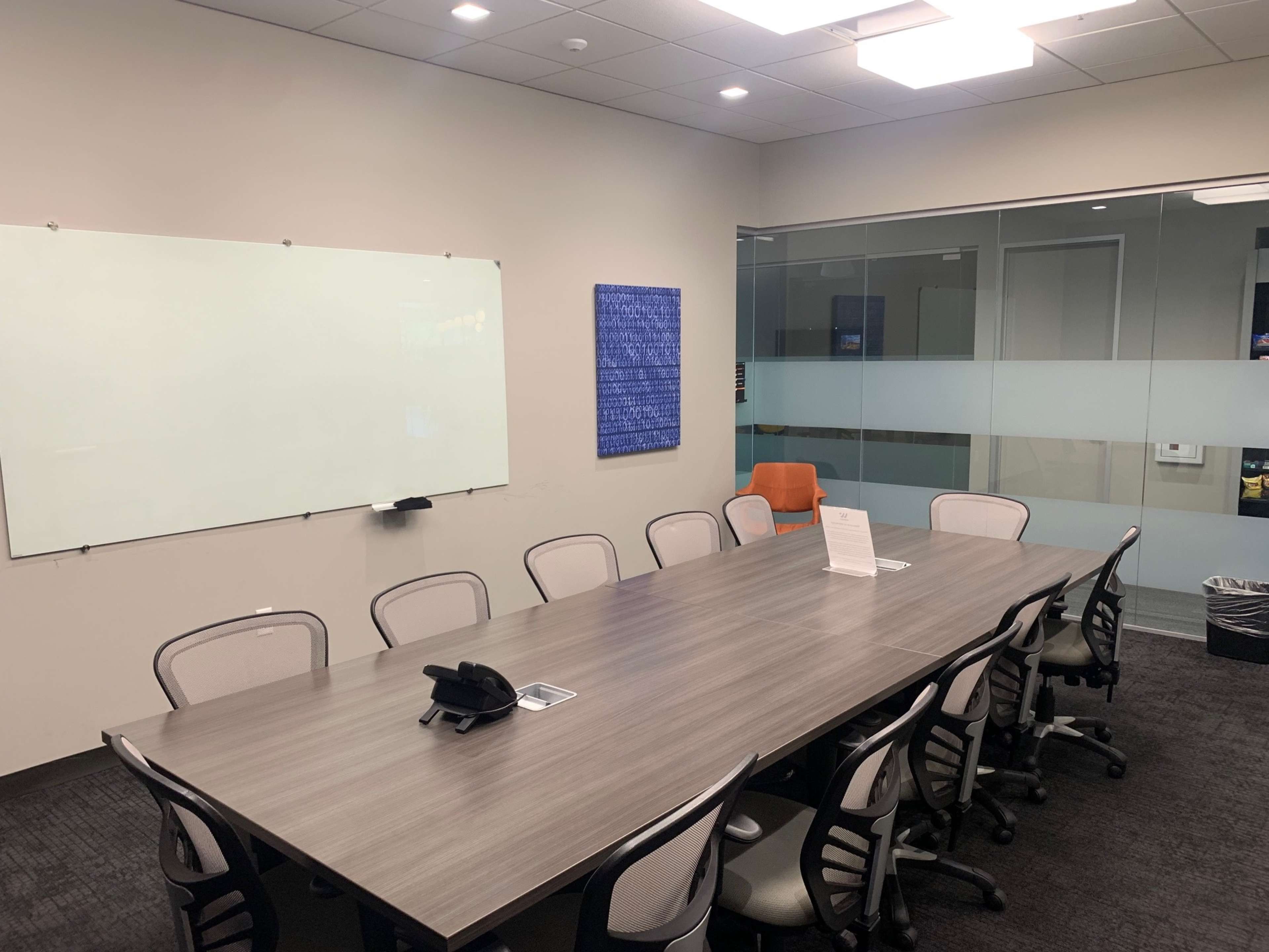 A conference room features a long table surrounded by ergonomic chairs, a whiteboard on one wall, and a glass partition revealing a workspace area.