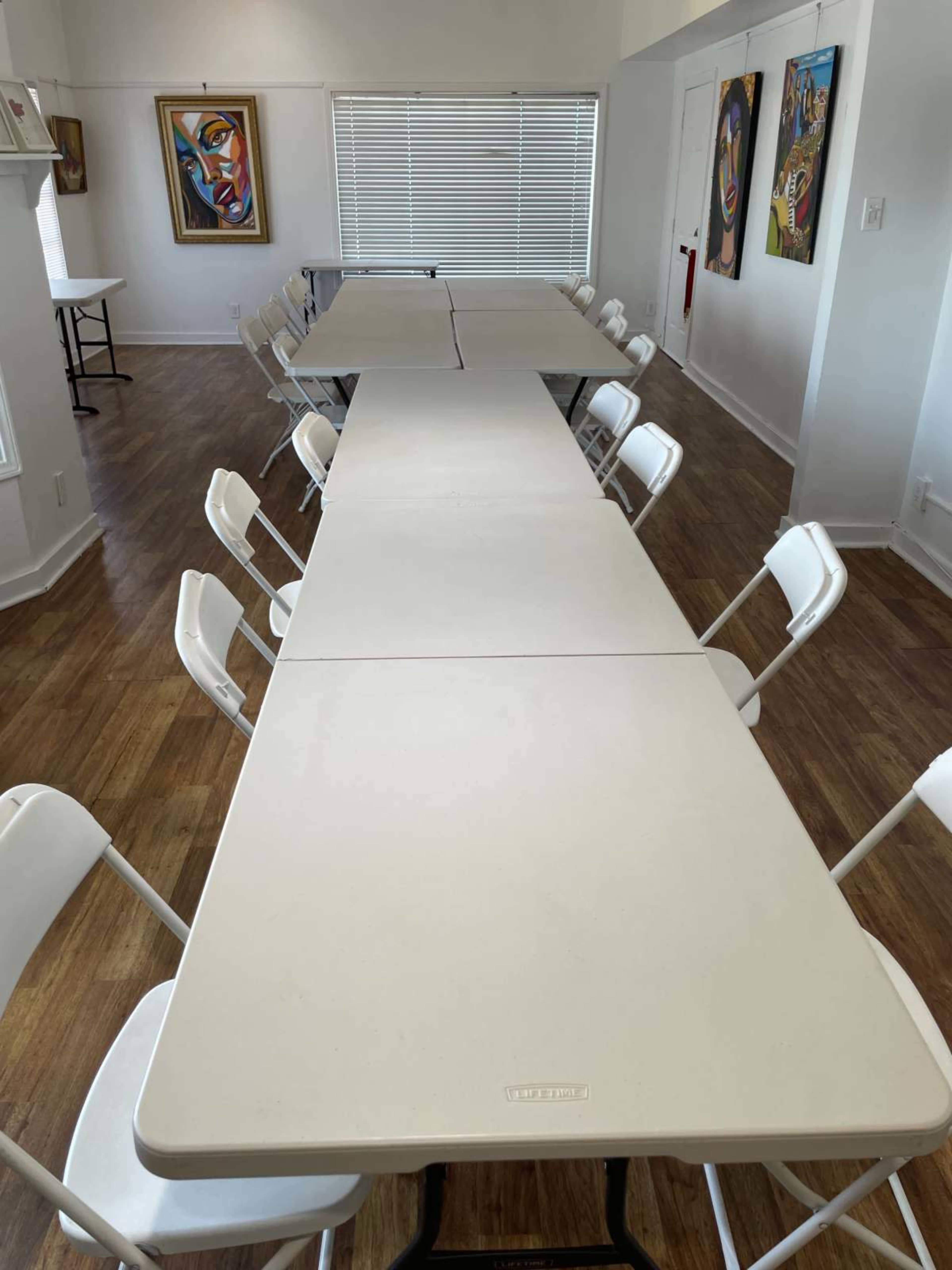 The image shows a long arrangement of white folding tables with white chairs on either side in a well-lit room featuring artwork on the walls.