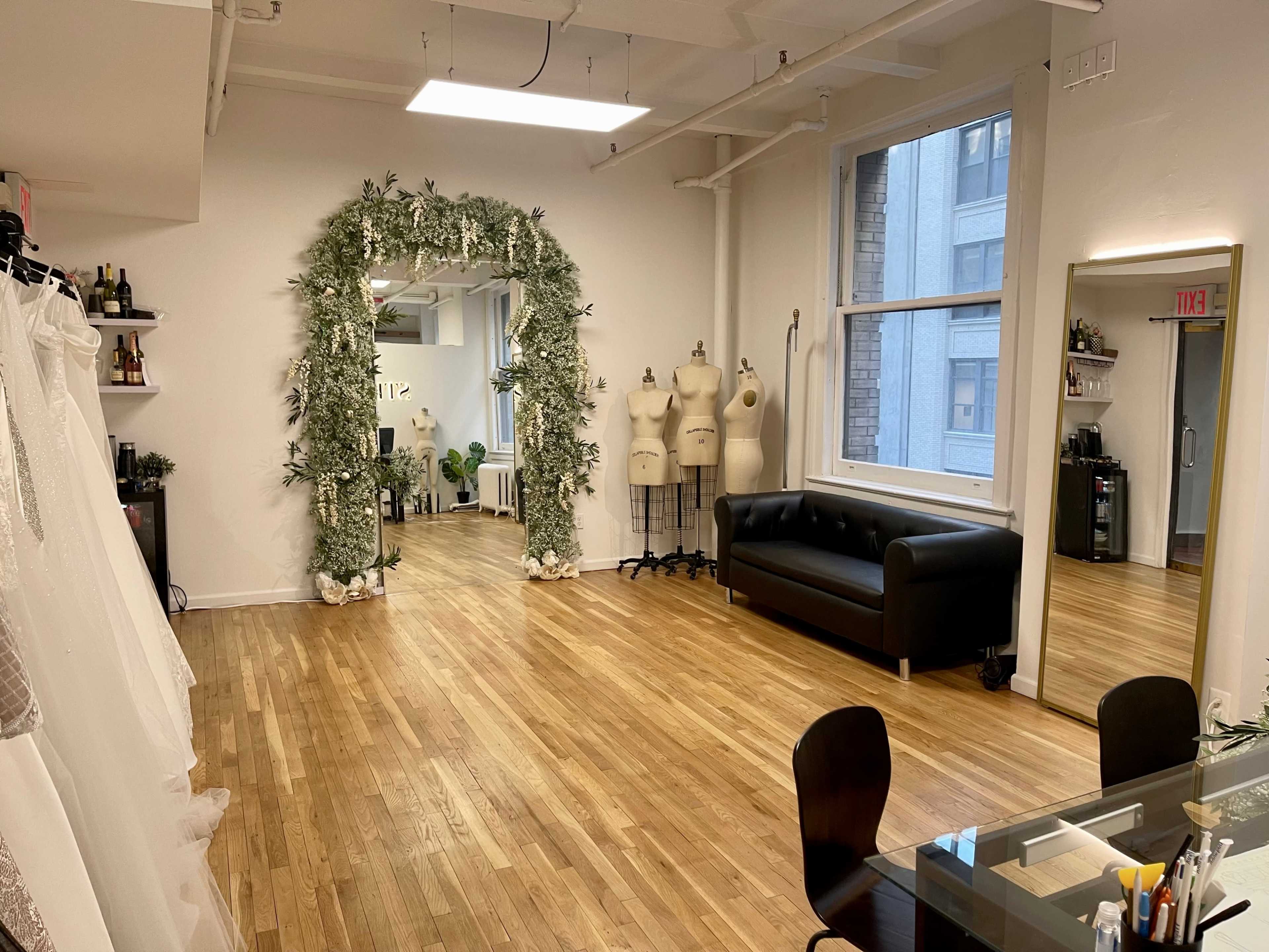 The image shows a spacious, well-lit room with a floral arch, a black sofa, dress-making mannequins, and a mirror along with a wooden floor.