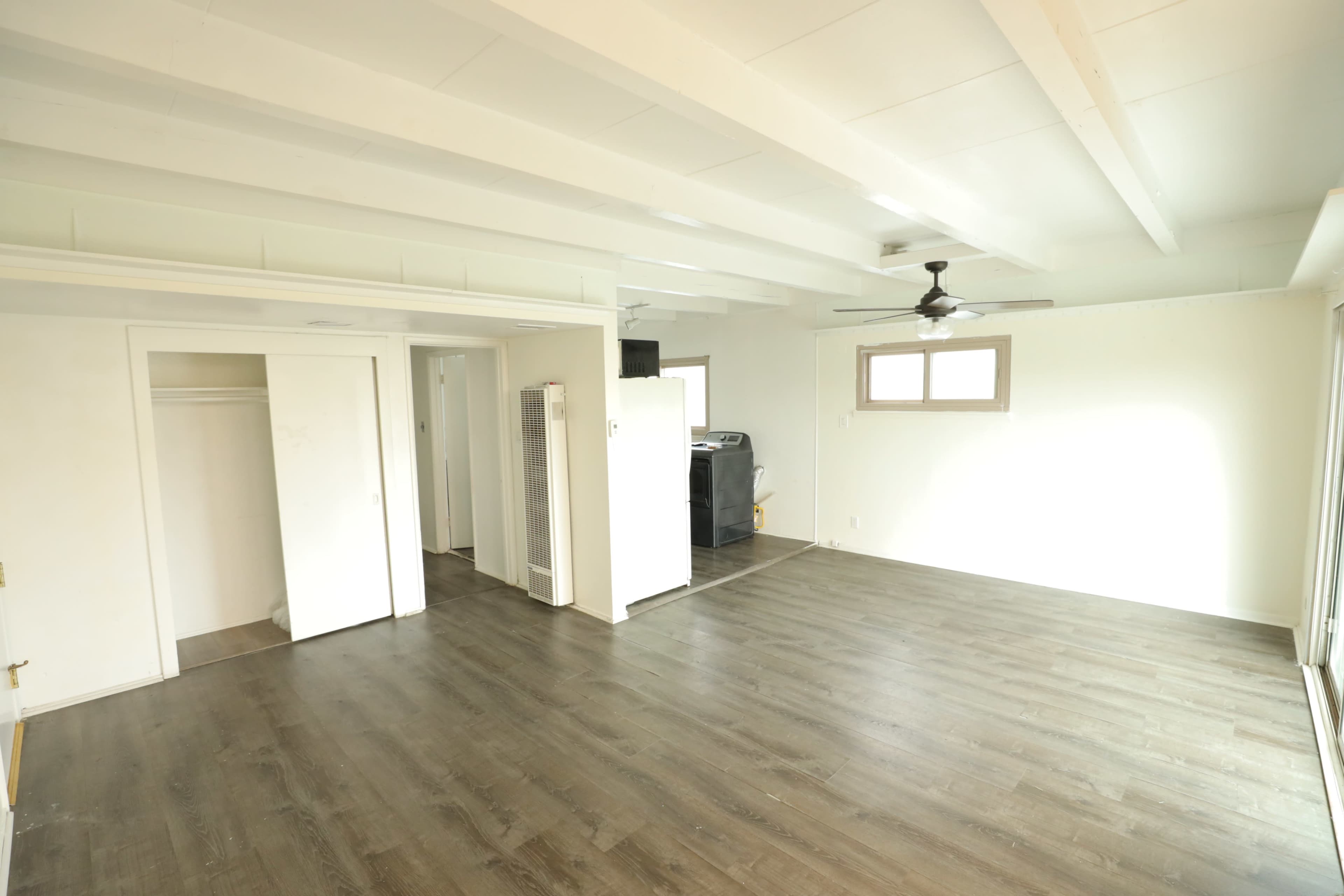 The image shows a spacious, empty room with wooden flooring, exposed beams on the ceiling, and minimal furnishings, leading to an open kitchen area.