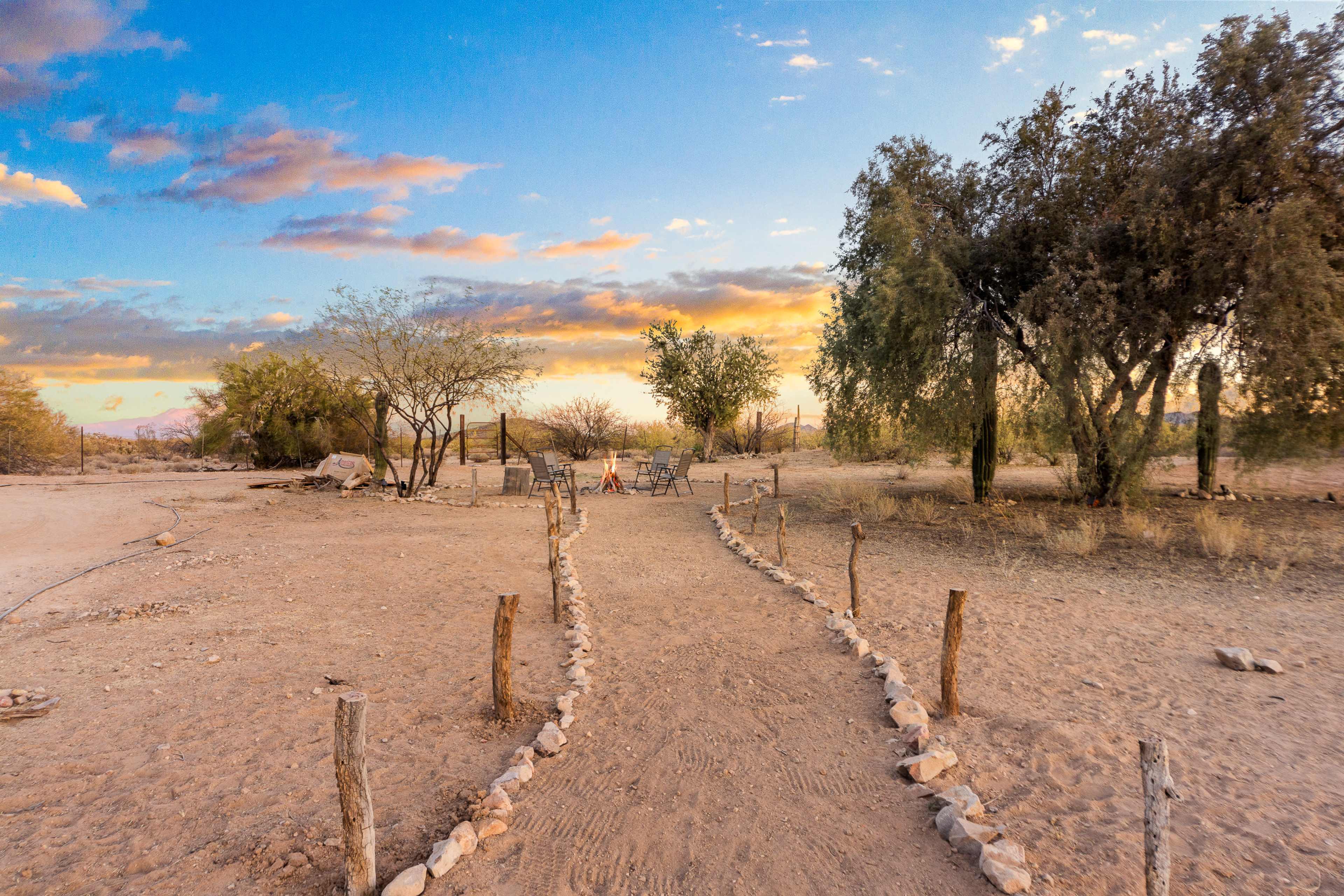 A dirt path lined with stones leads to a fire pit surrounded by chairs in a desert landscape at sunset.