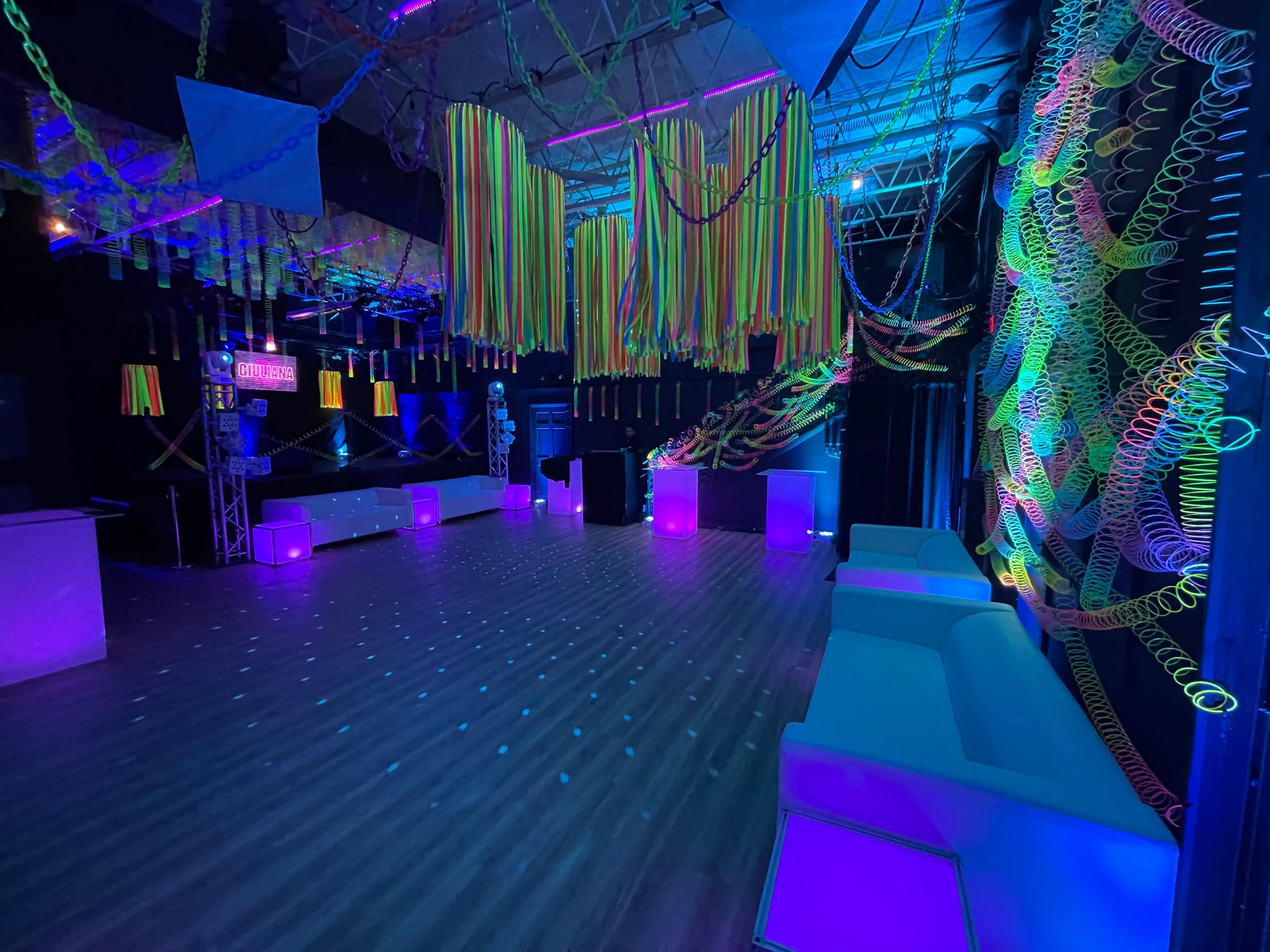 A dimly lit event space decorated with colorful streamers and neon accents, featuring white sofas and illuminated tables.