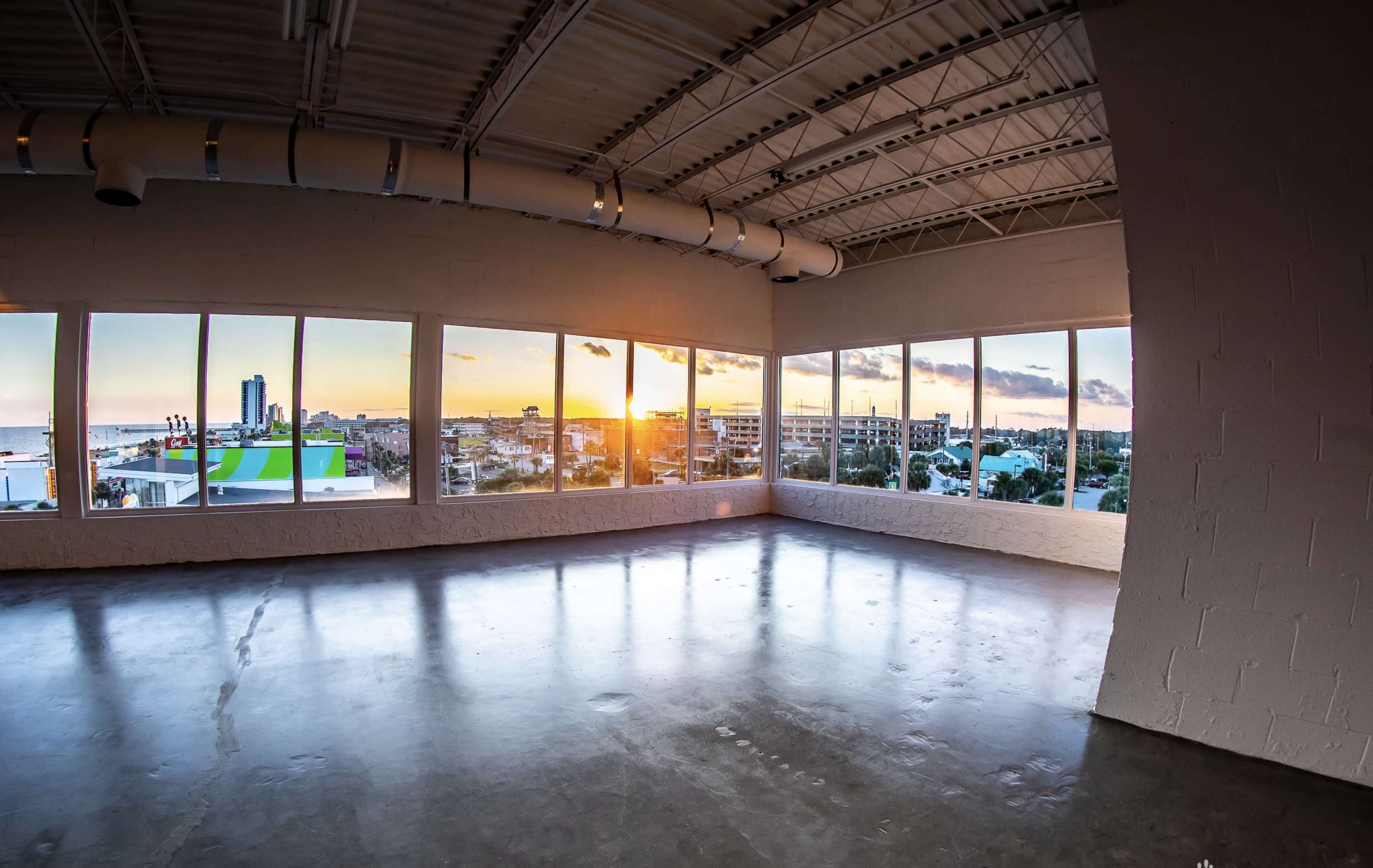 A vacant room with large windows offers a view of a sunset over a cityscape.