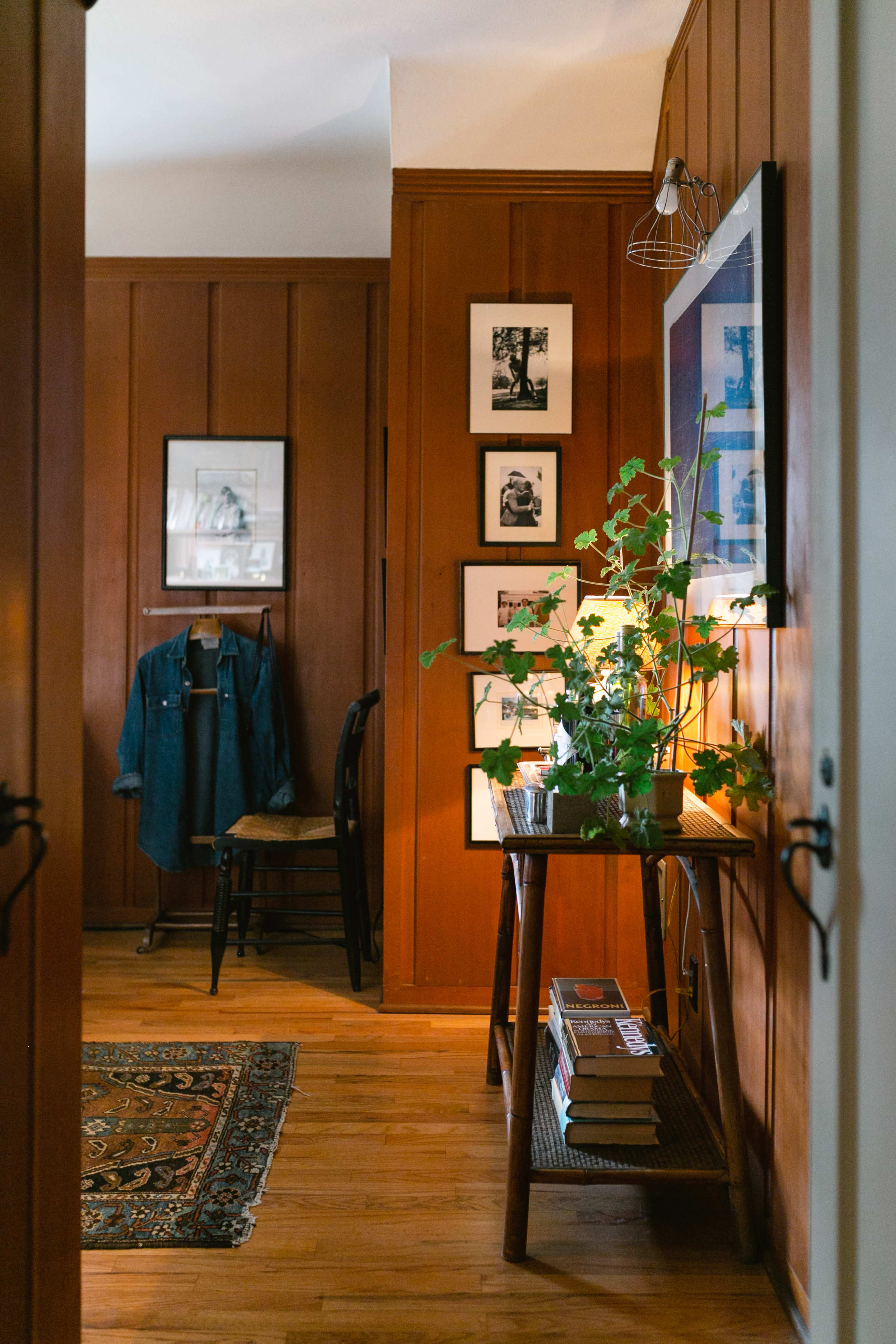 A hallway features wooden paneled walls, a small table with plants and books, and framed photographs hanging nearby.