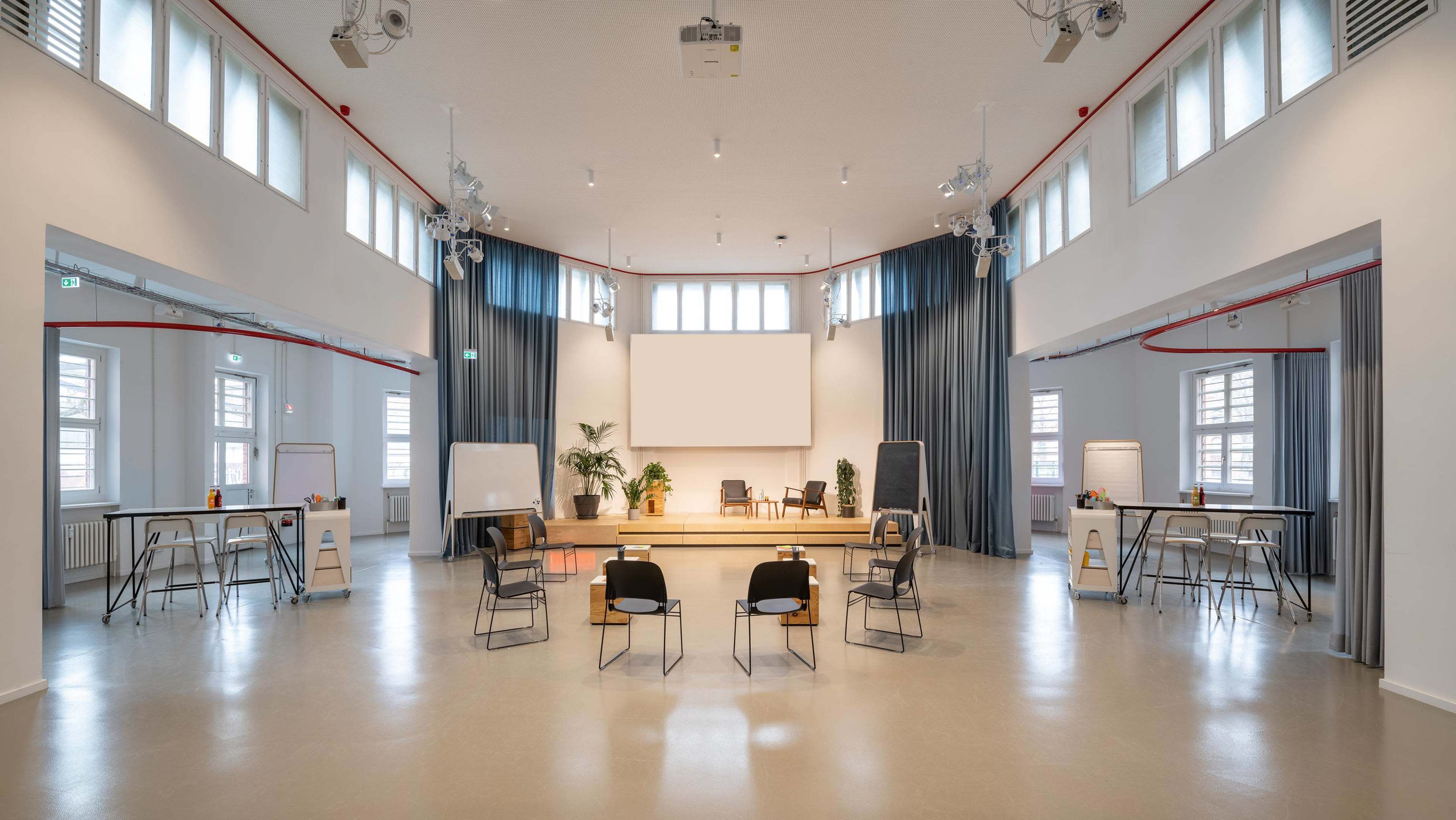 The image shows a spacious, well-lit seminar room with a large empty screen, seating arranged in a circle, and several high tables along the sides.