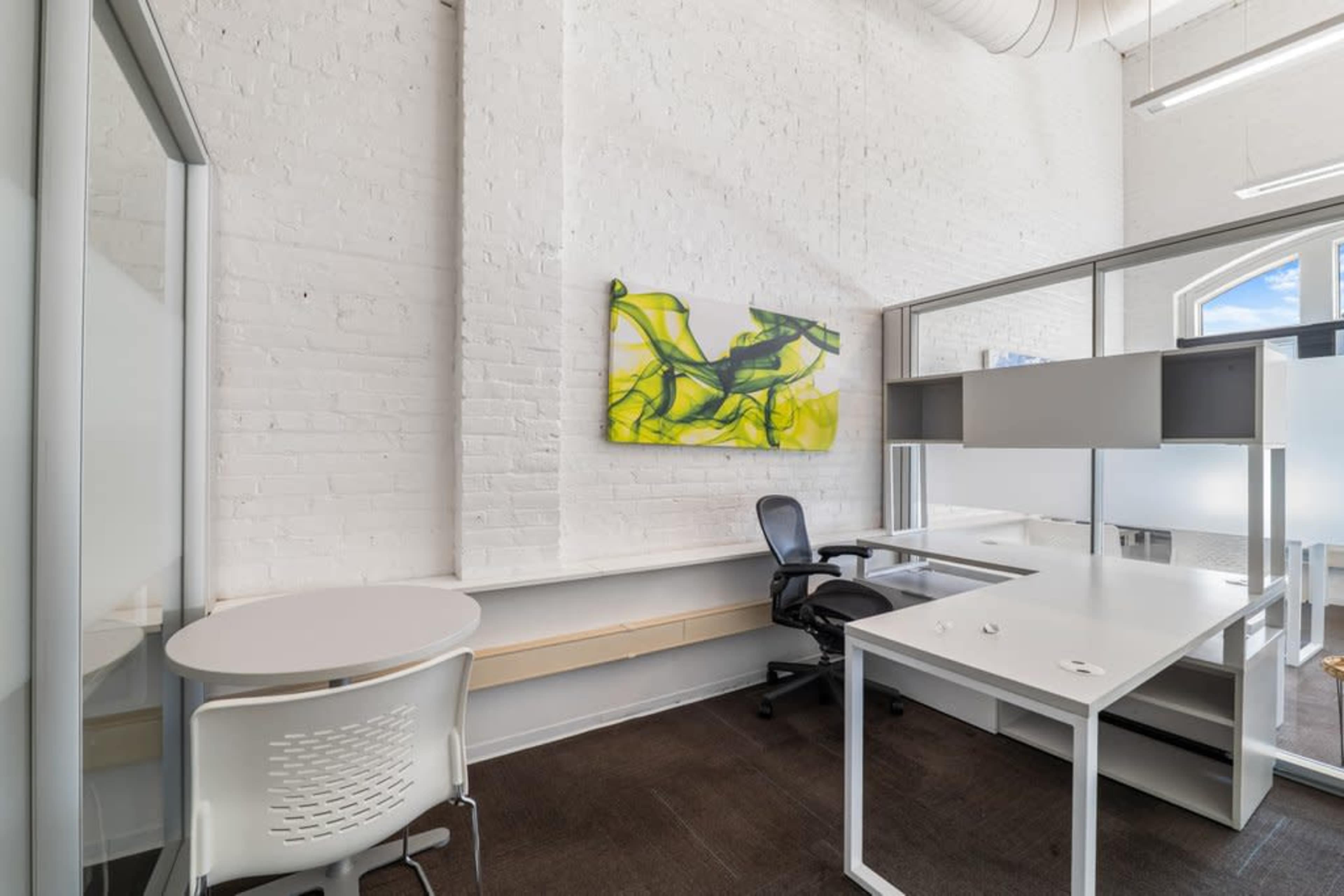 The image shows a bright office space featuring a white brick wall, a colorful abstract painting, and modern furniture including a desk, chair, and a small table.