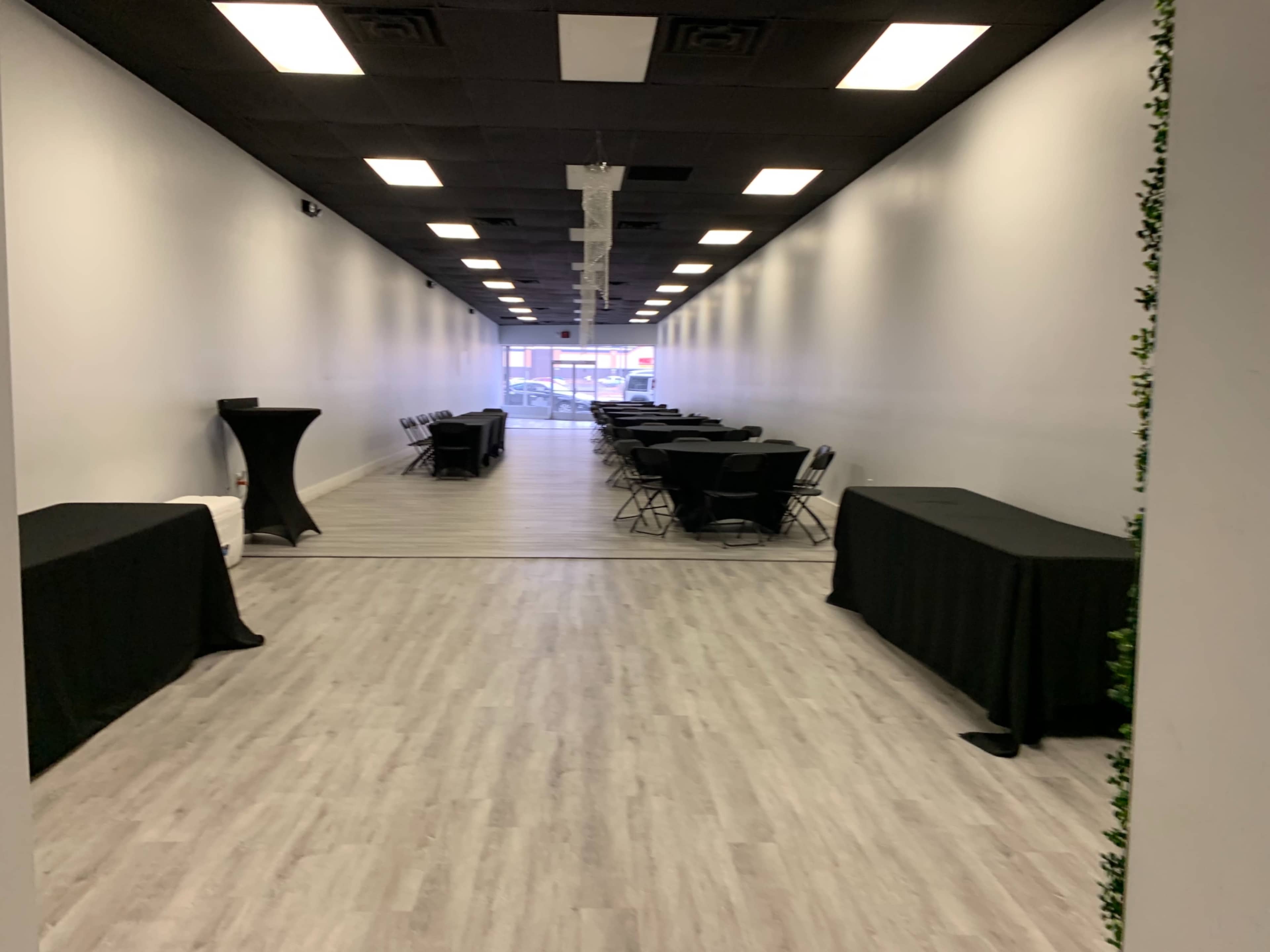 A long, narrow hallway with black tables and chairs on either side, leading to a brightly lit entrance at the far end.