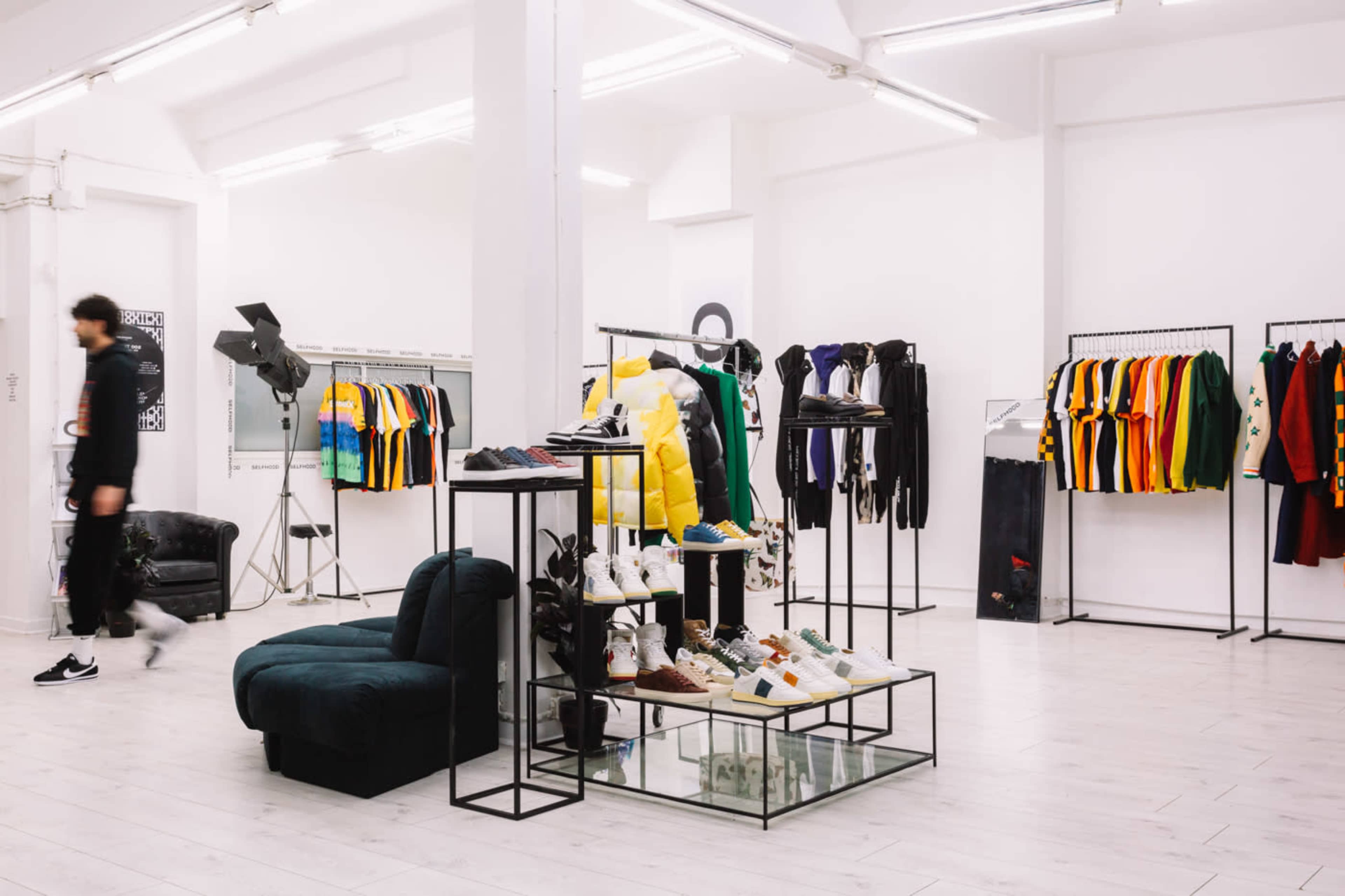The image shows a modern clothing store with racks of colorful apparel and shoes displayed on glass tables, while a person is walking in the background.