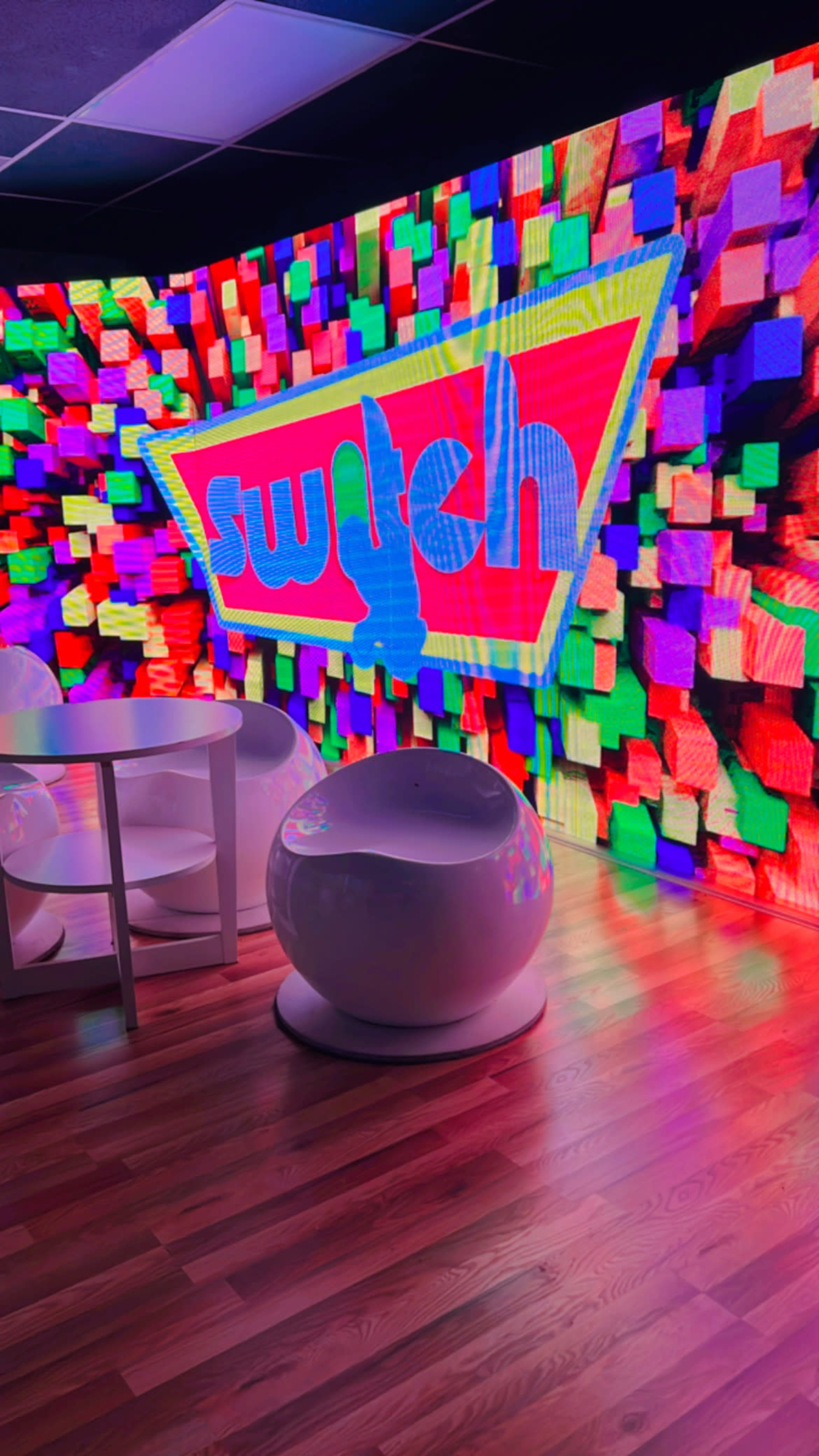 The image shows a vibrant, colorful wall display with the word "switch" illuminated, accompanied by modern, white furniture in a room.