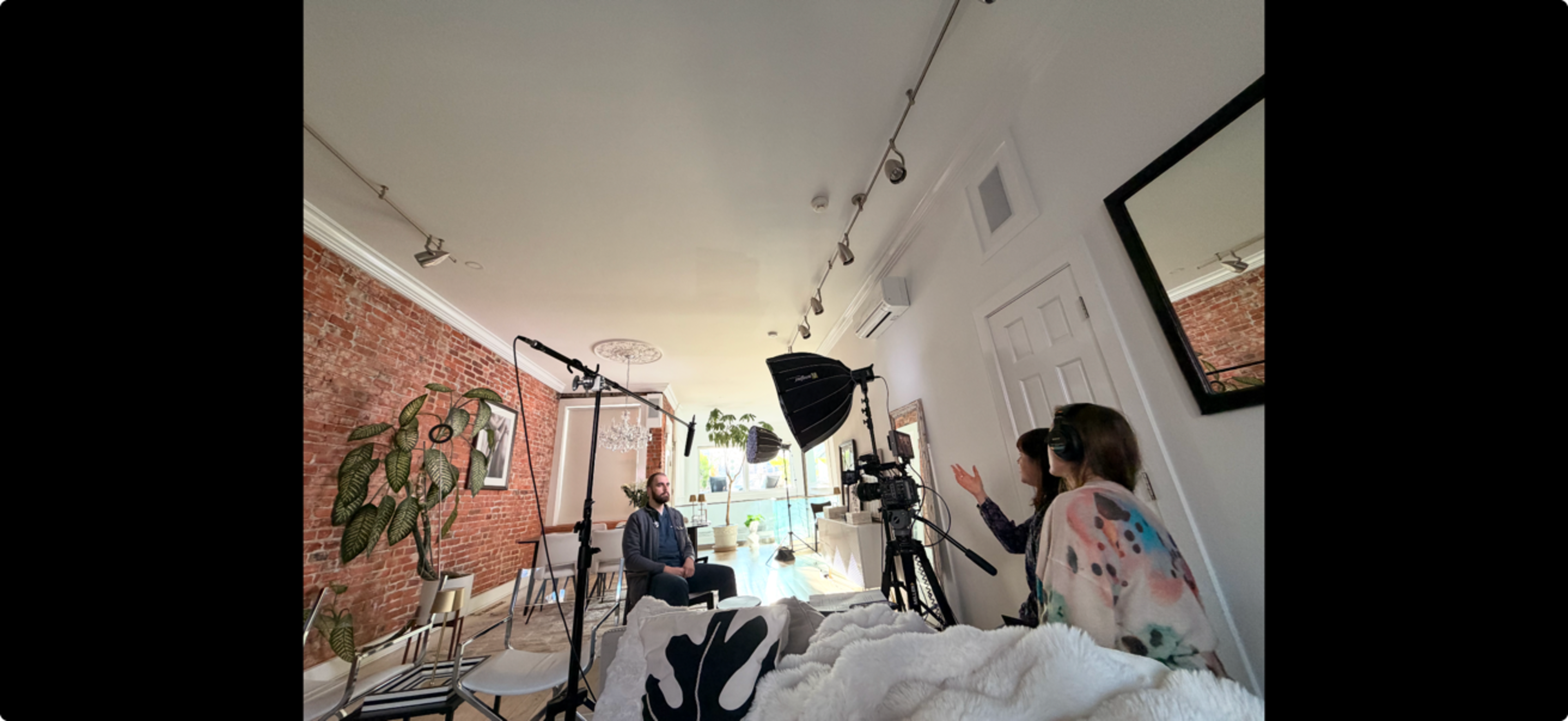 A man sits in a bright room with exposed brick walls while two people, one seated and the other behind a camera, engage in a discussion.