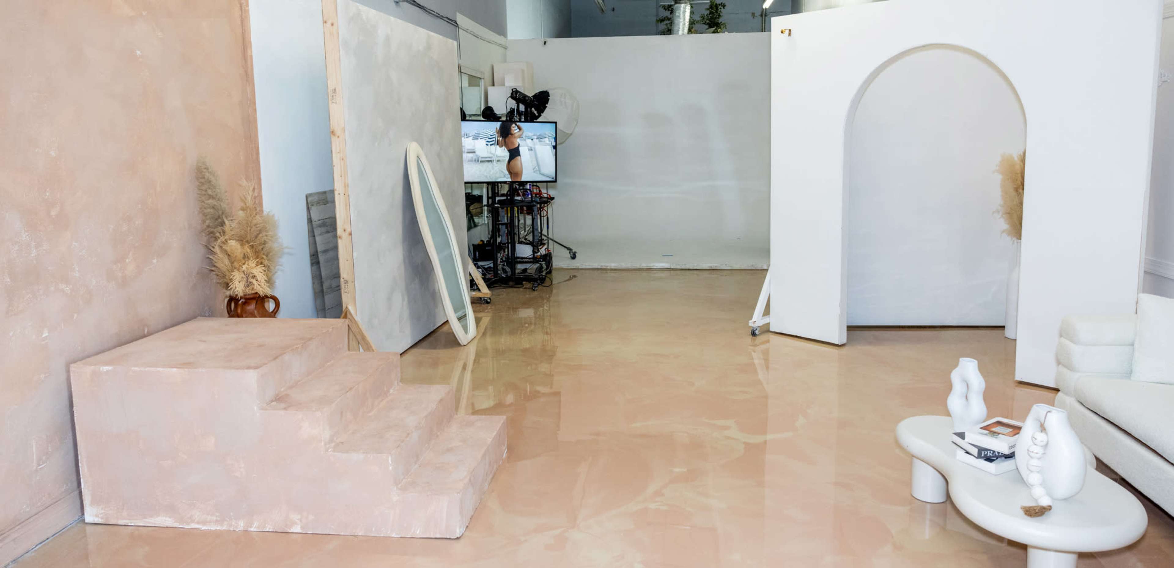 The image shows a minimalist interior space with a light-colored floor, a staircase made of a textured material, and a large curved archway, along with a television displaying a video in the background.