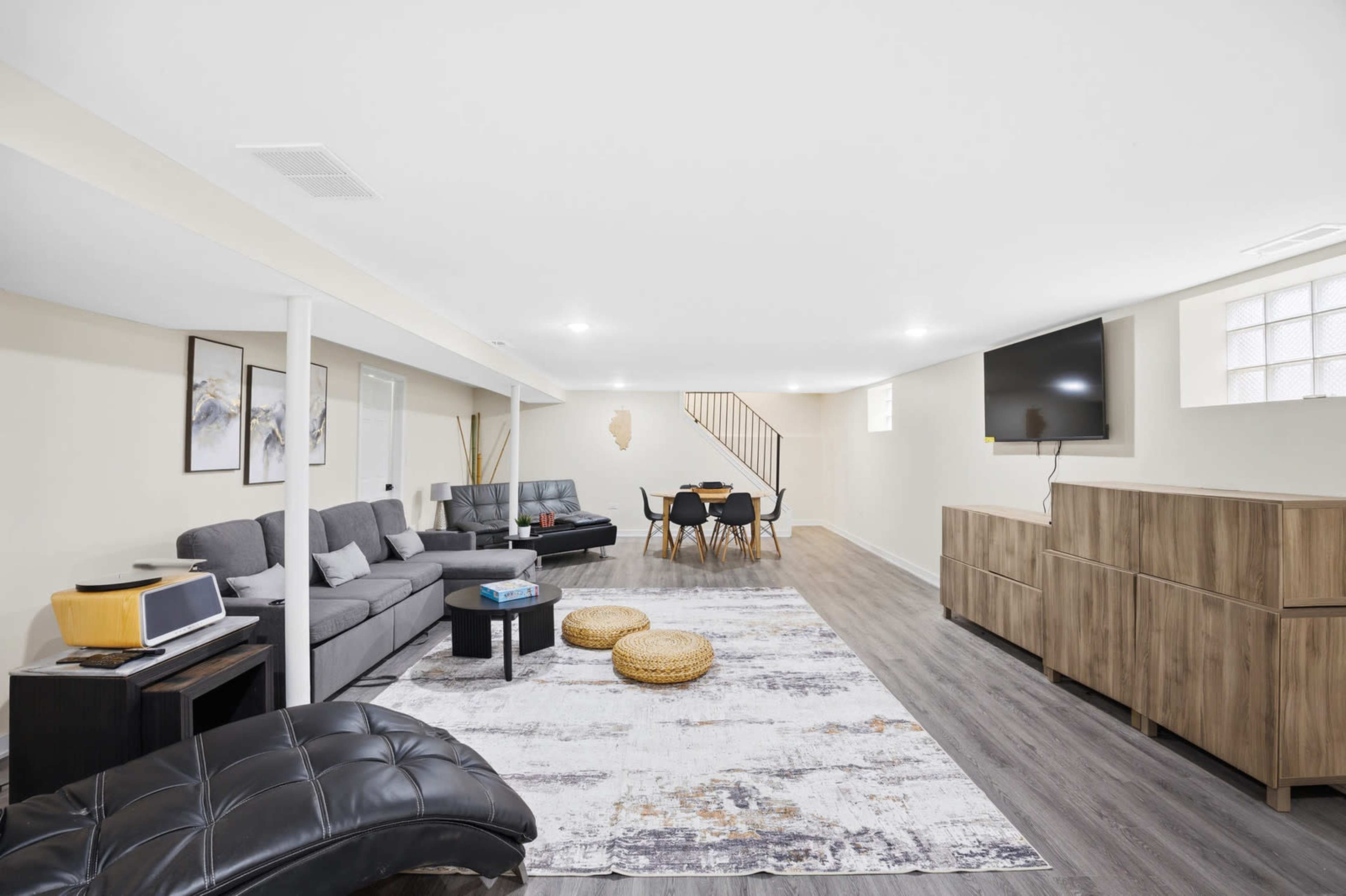 The image shows a spacious, well-lit basement furnished with a gray sectional sofa, a dining table, and light-colored walls.