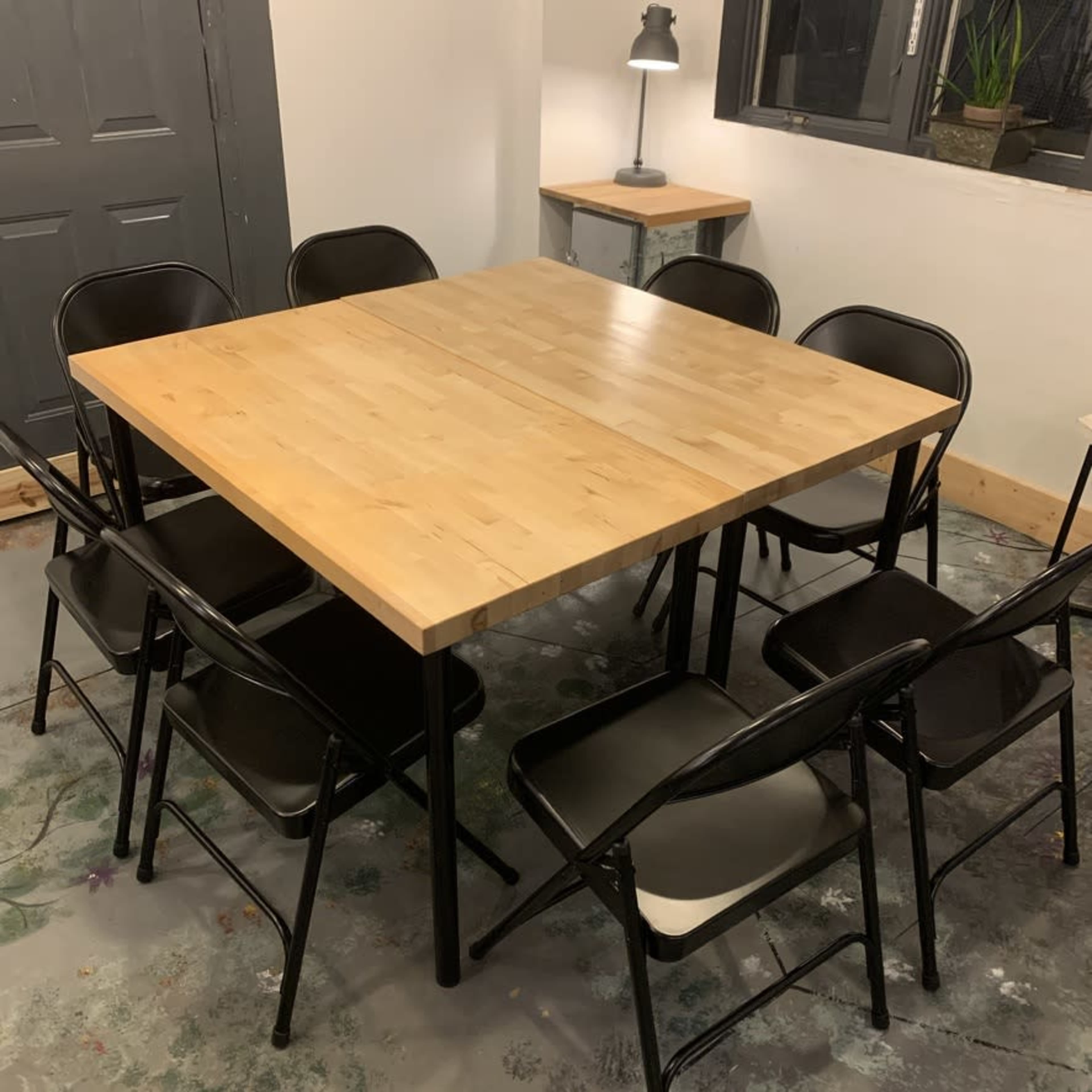 A square wooden table with a light-colored surface is surrounded by eight black folding chairs in a well-lit room.