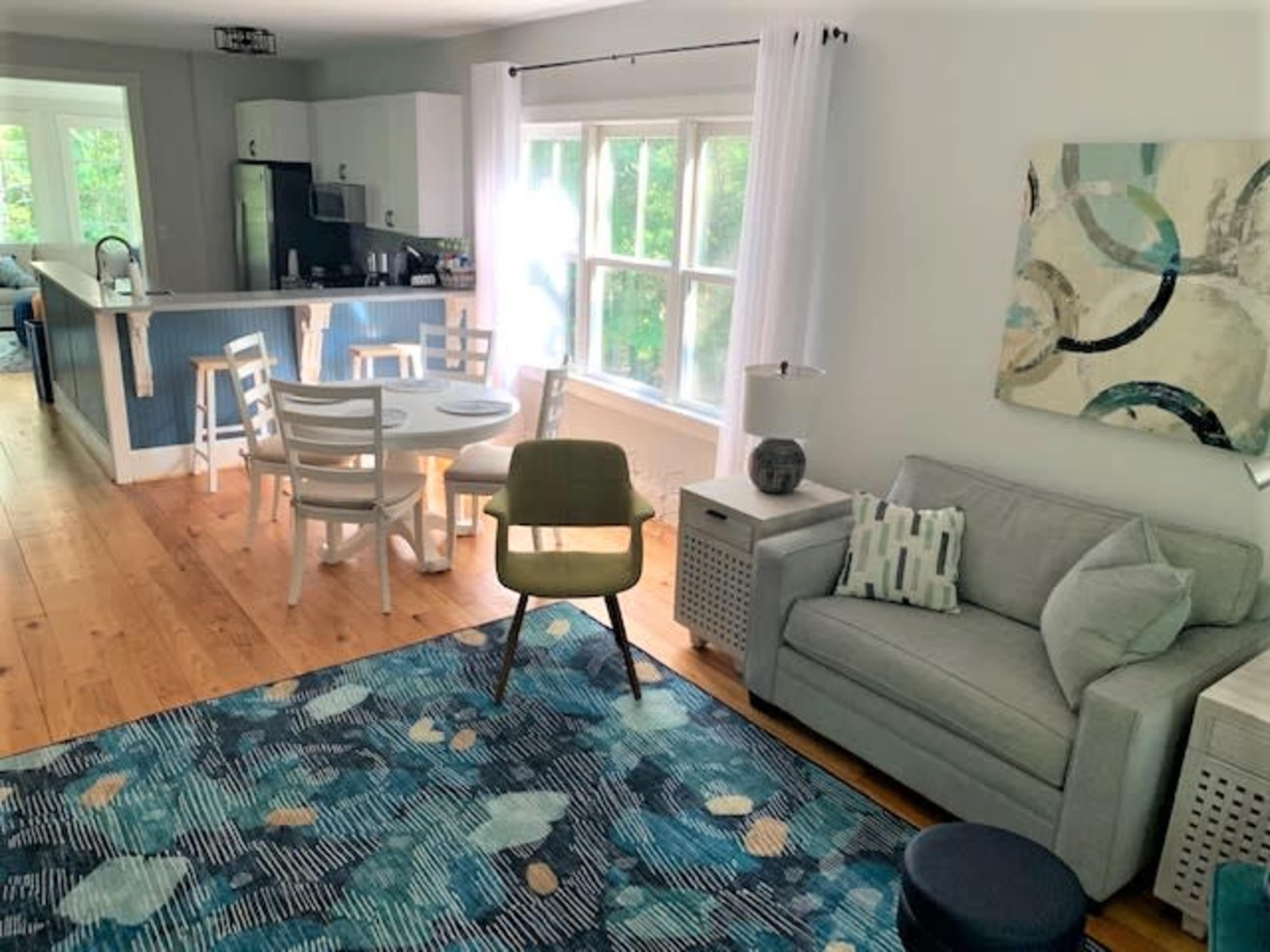 The image shows a bright, open living space featuring a light gray sofa, a colorful area rug, a dining table, and a kitchen area with white cabinetry and blue accents.