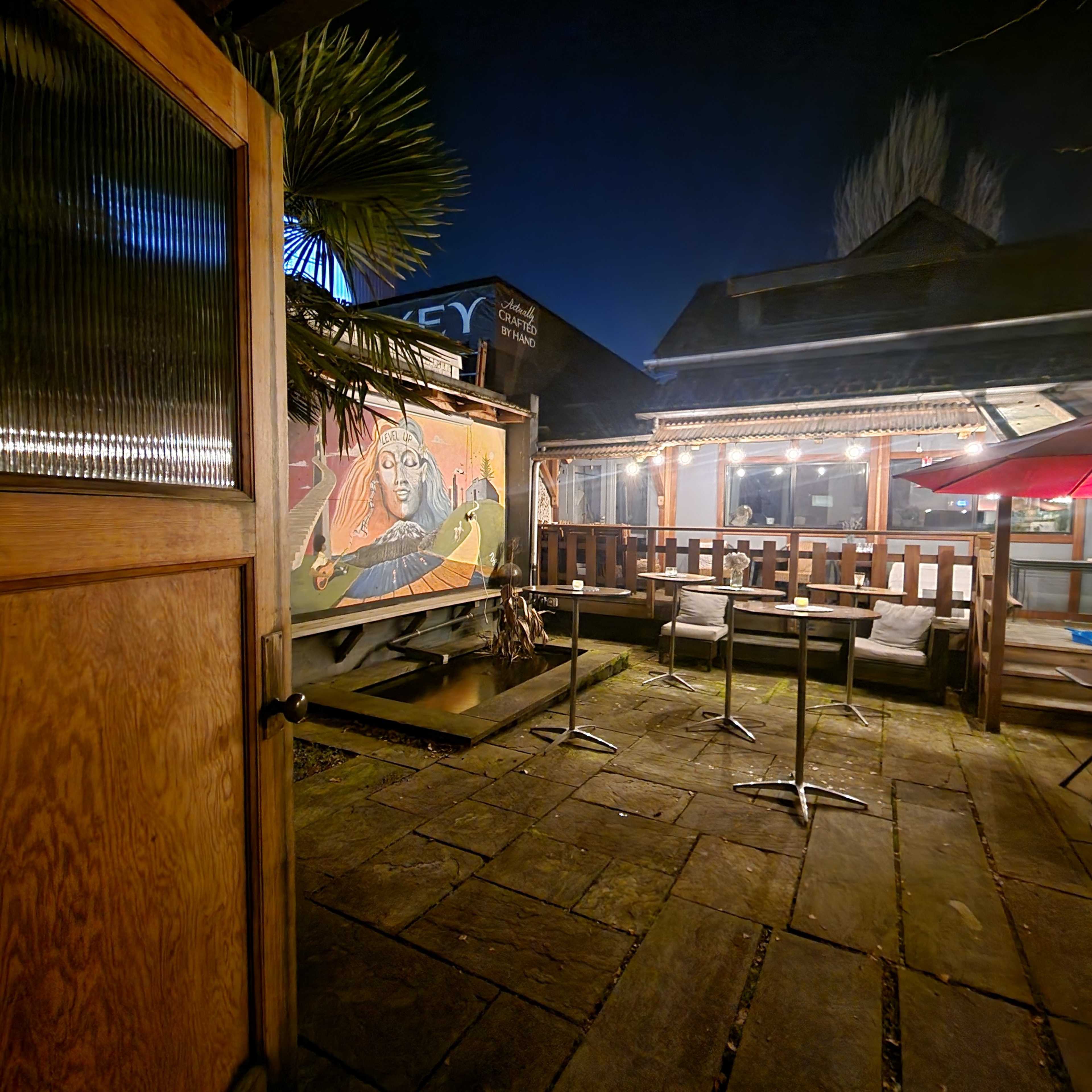 A wooden door opens to reveal a partially illuminated outdoor patio with a mural and several empty chairs under string lights.