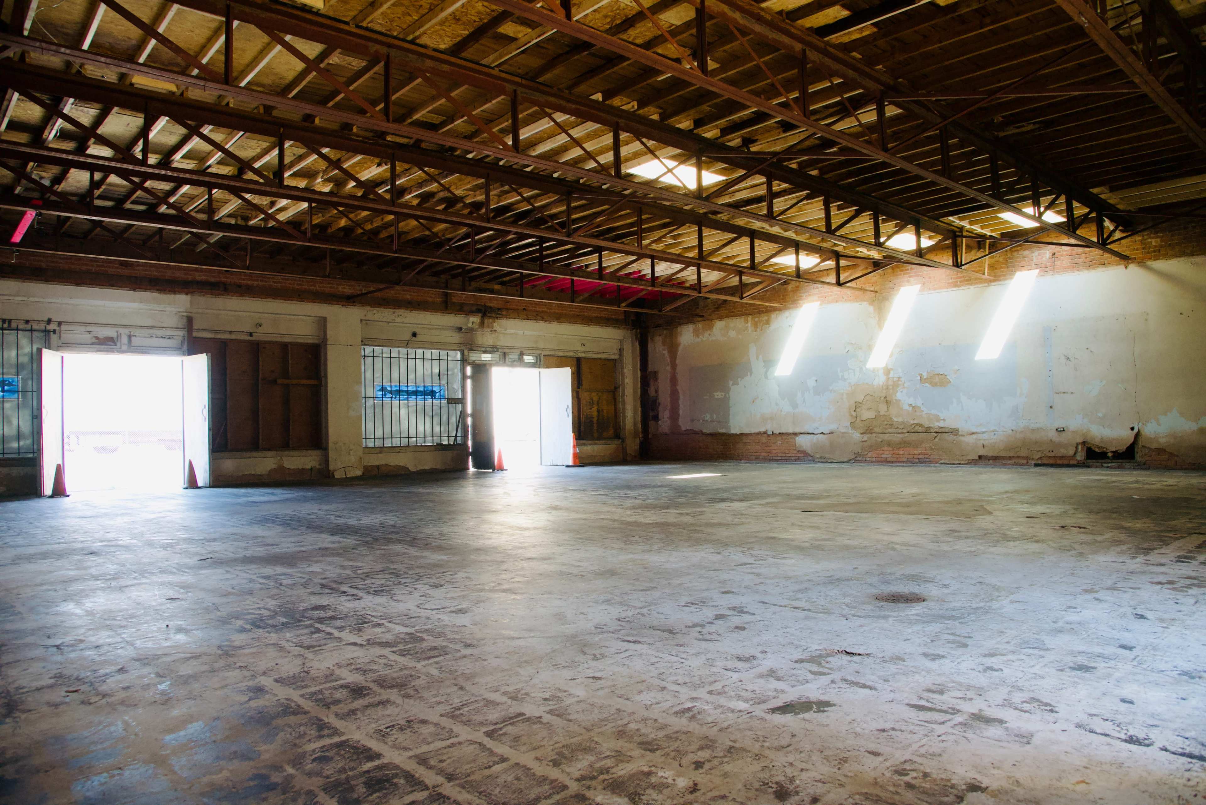 An empty, industrial space with exposed beams, concrete flooring, and light streaming through windows on the walls.