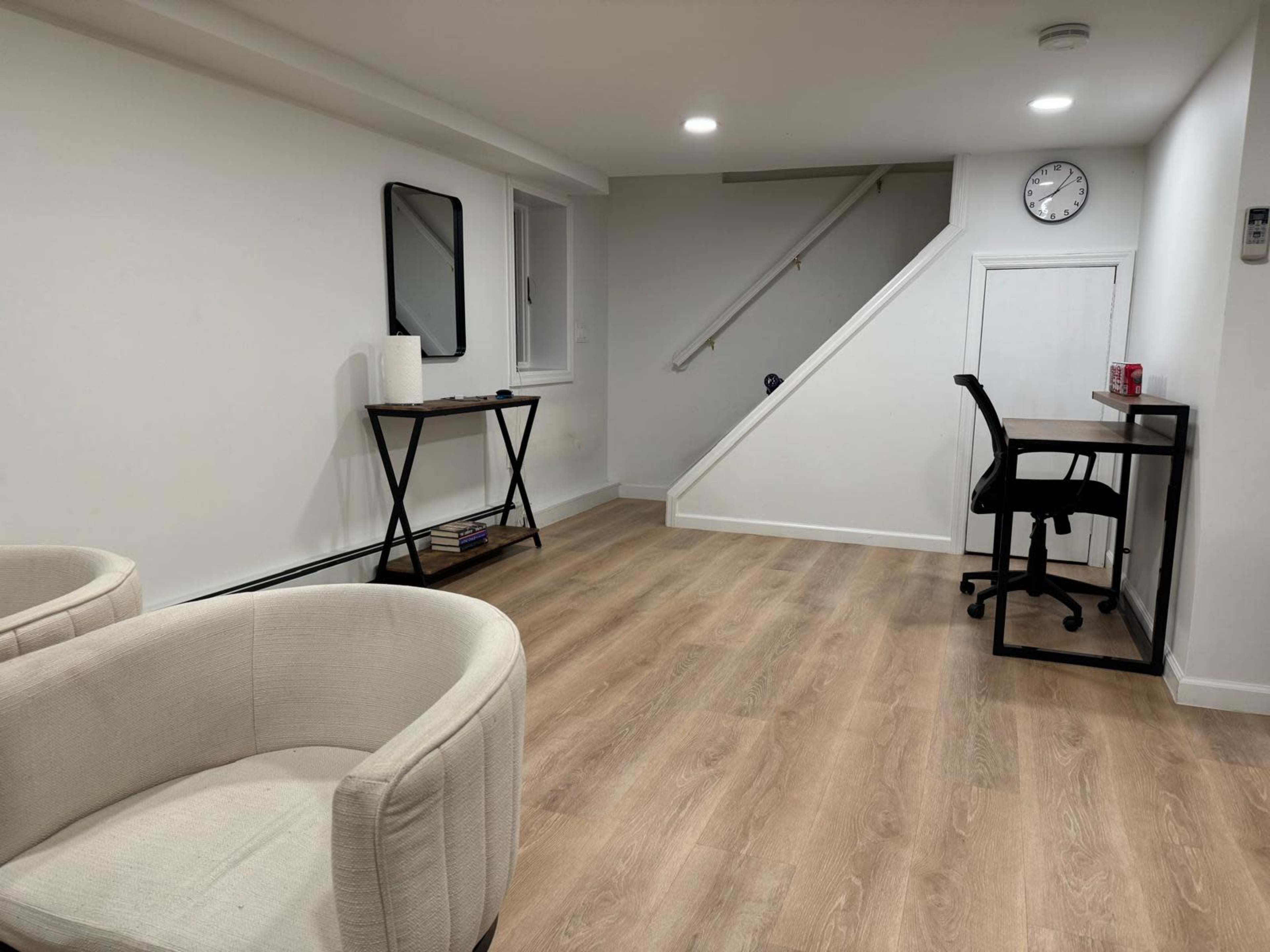 The image shows a minimally furnished, bright basement room with two chairs, a small table, a clock on the wall, and a staircase leading to another level.