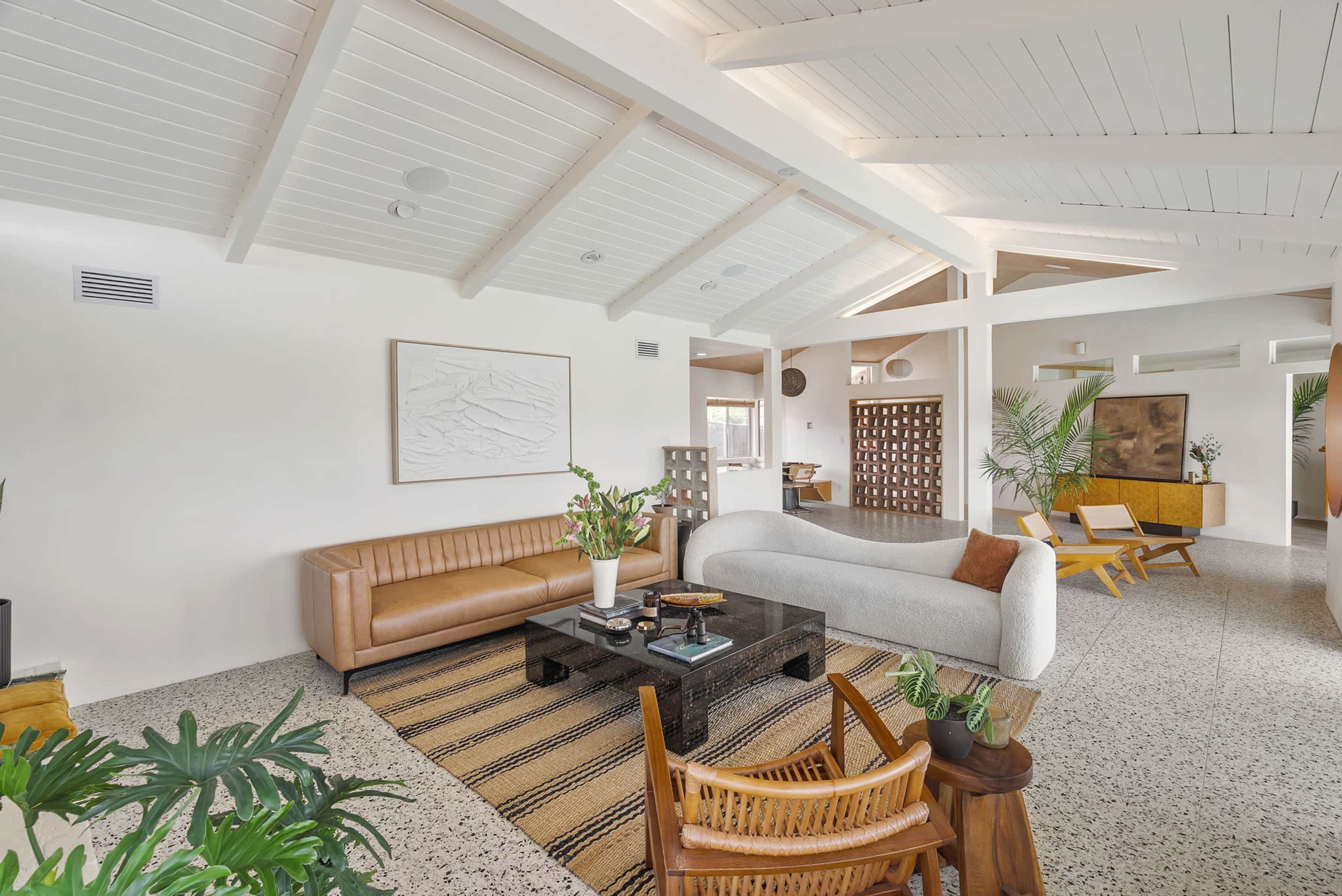 The image shows a spacious living area with a brown leather sofa, a white fabric couch, and various seating arrangements, featuring a patterned rug and indoor plants.