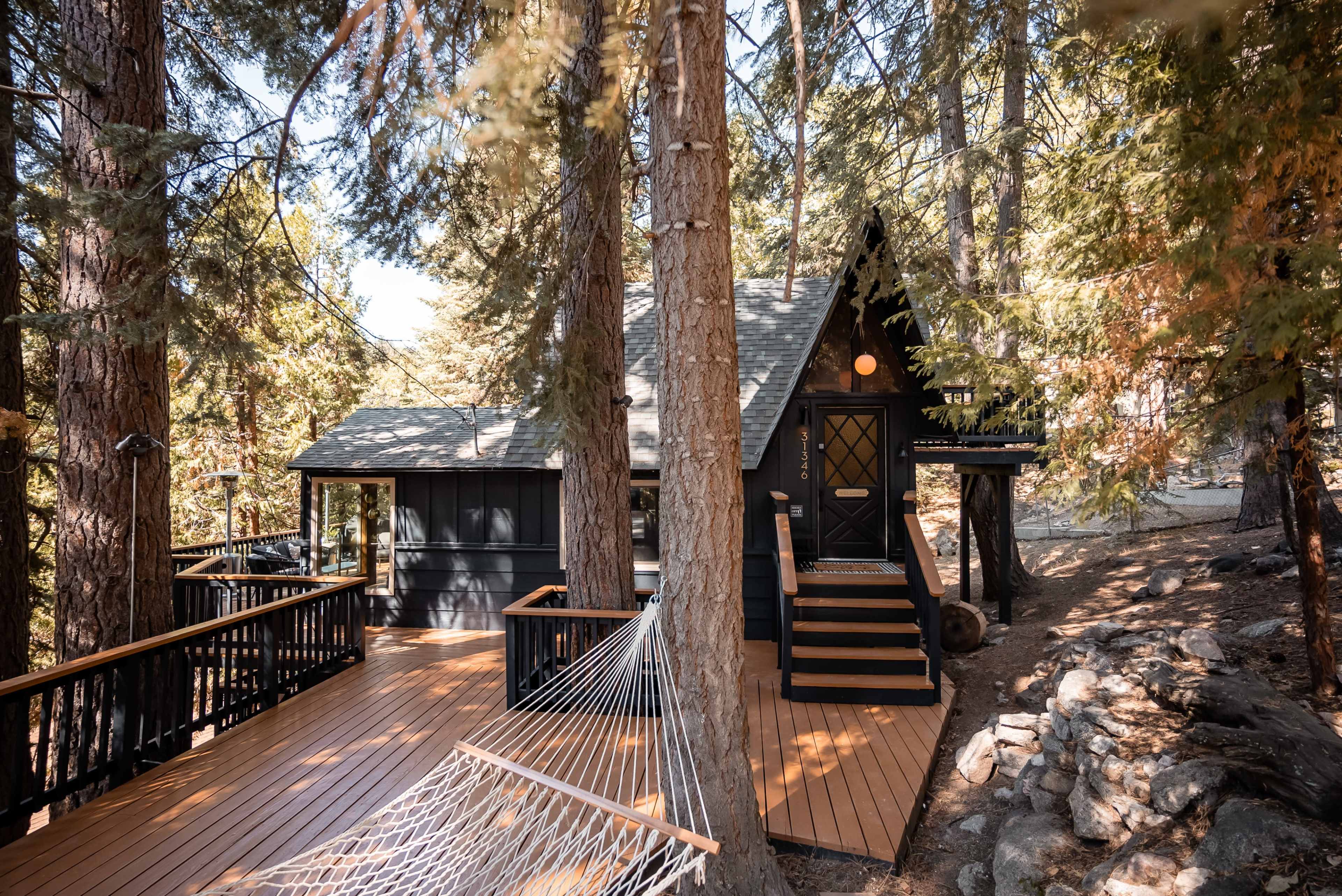 A black chalet-style cabin with a large wooden deck and hammock is surrounded by tall trees in a forested area.