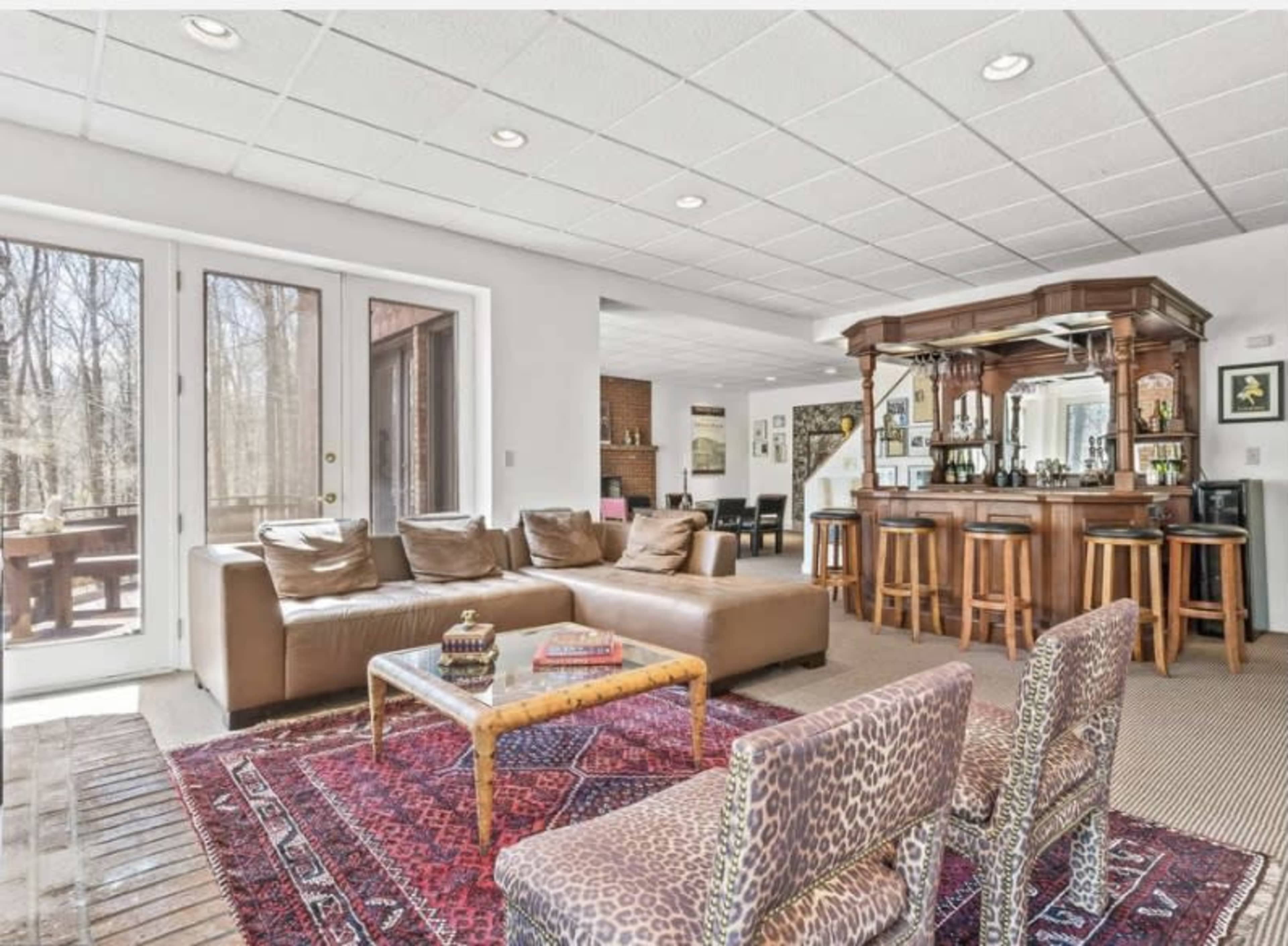 The image shows a spacious living area with a bar, seating on a patterned rug, and large windows overlooking a wooded view.