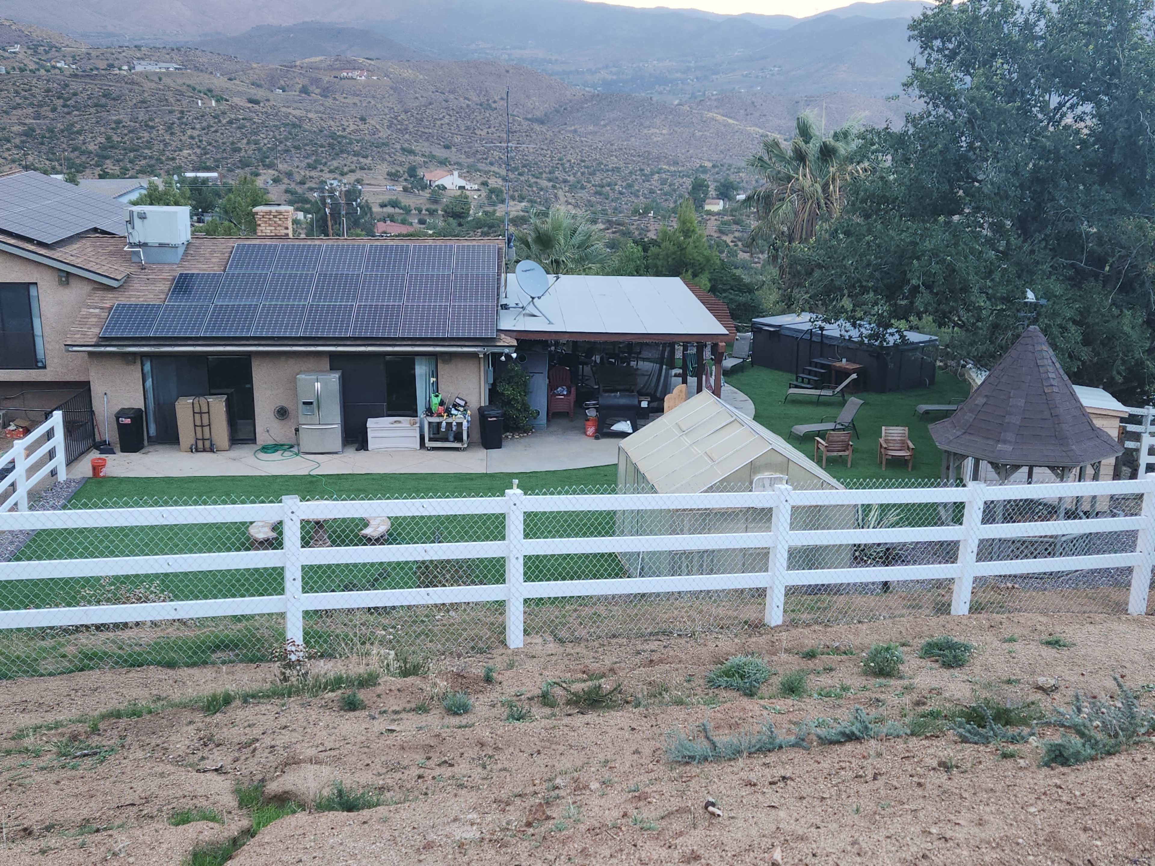 The image shows a hillside property featuring a house with solar panels, a patio area, an outdoor gazebo, and a small storage shed.