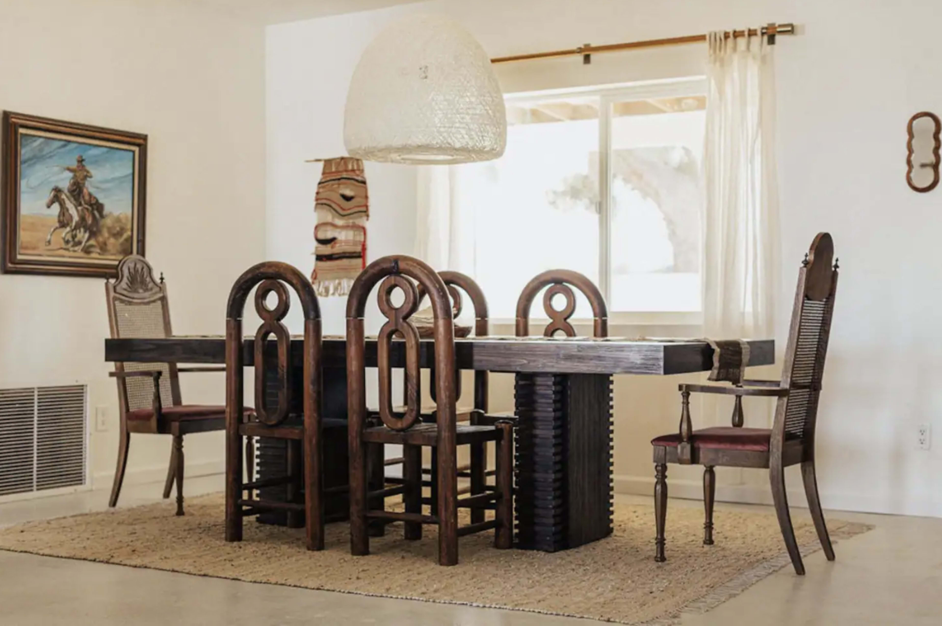 A dining room features a large wooden table surrounded by various styled chairs, with a woven light fixture overhead and a window letting in natural light.