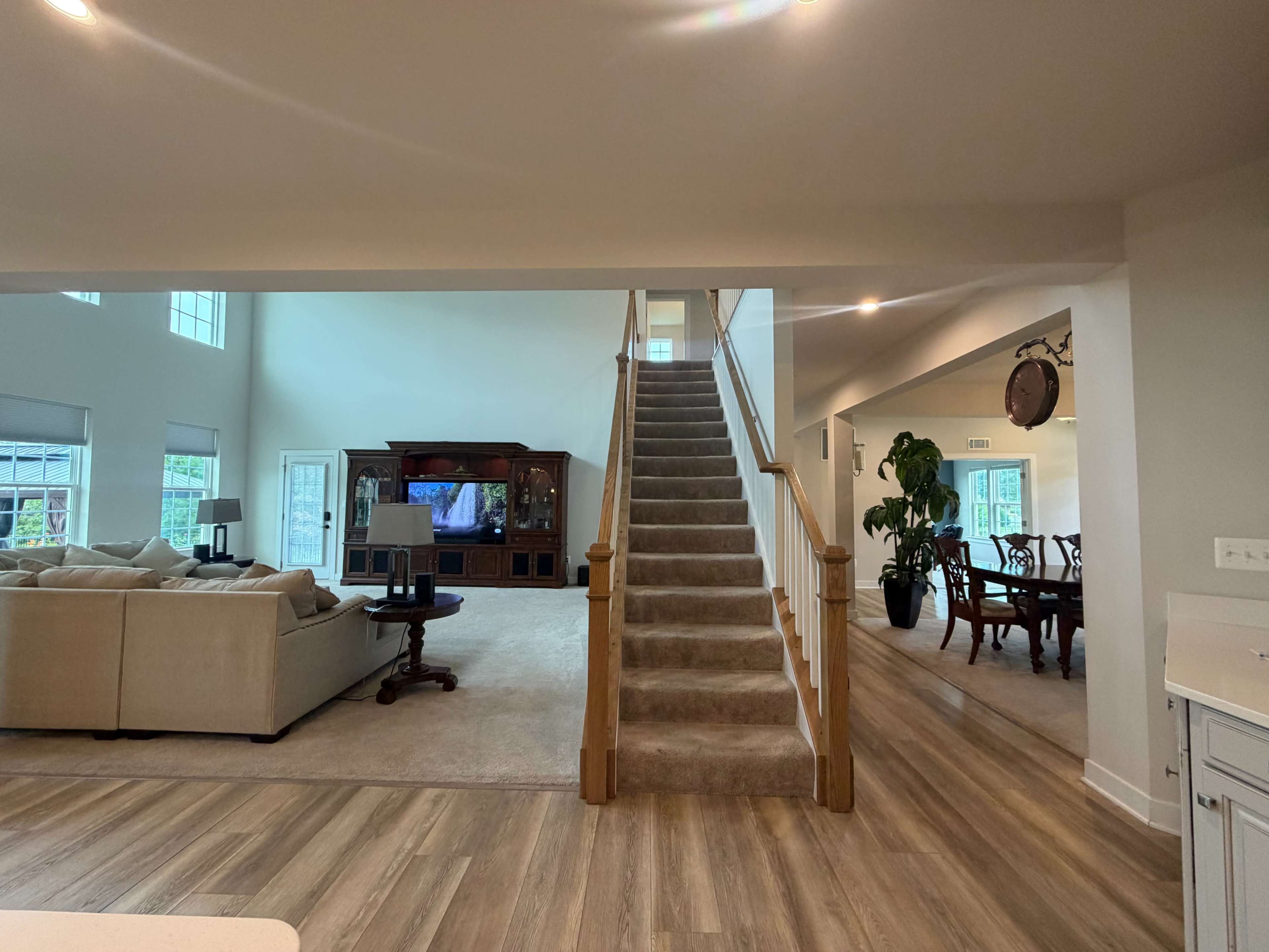 The image shows a spacious living area with a staircase leading to an upper level, flanked by a living room furnished with a sofa set and a dining area visible in the background.