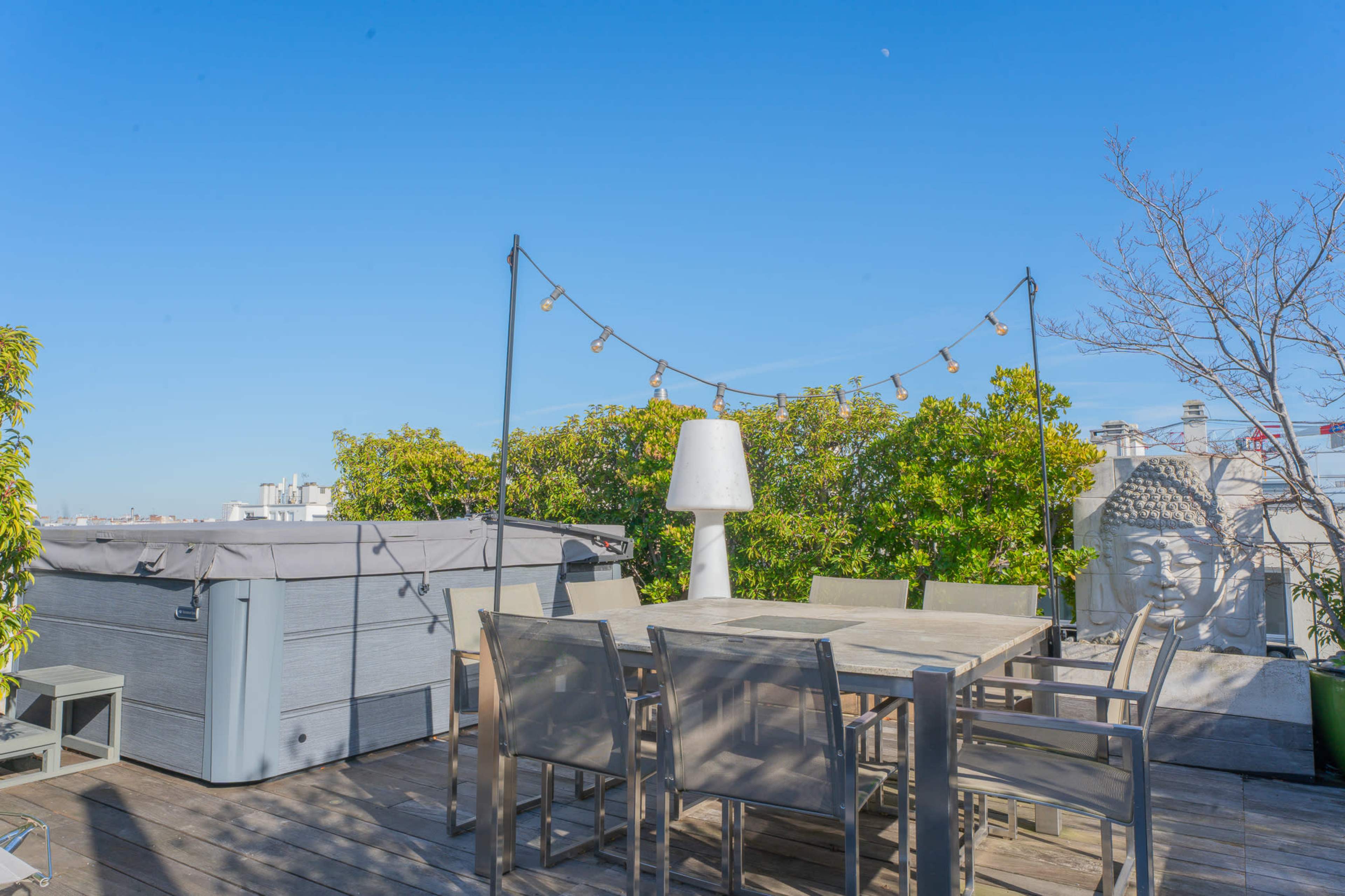 A rooftop terrace features a wooden table with several chairs, surrounded by greenery and a hot tub, along with string lights overhead.