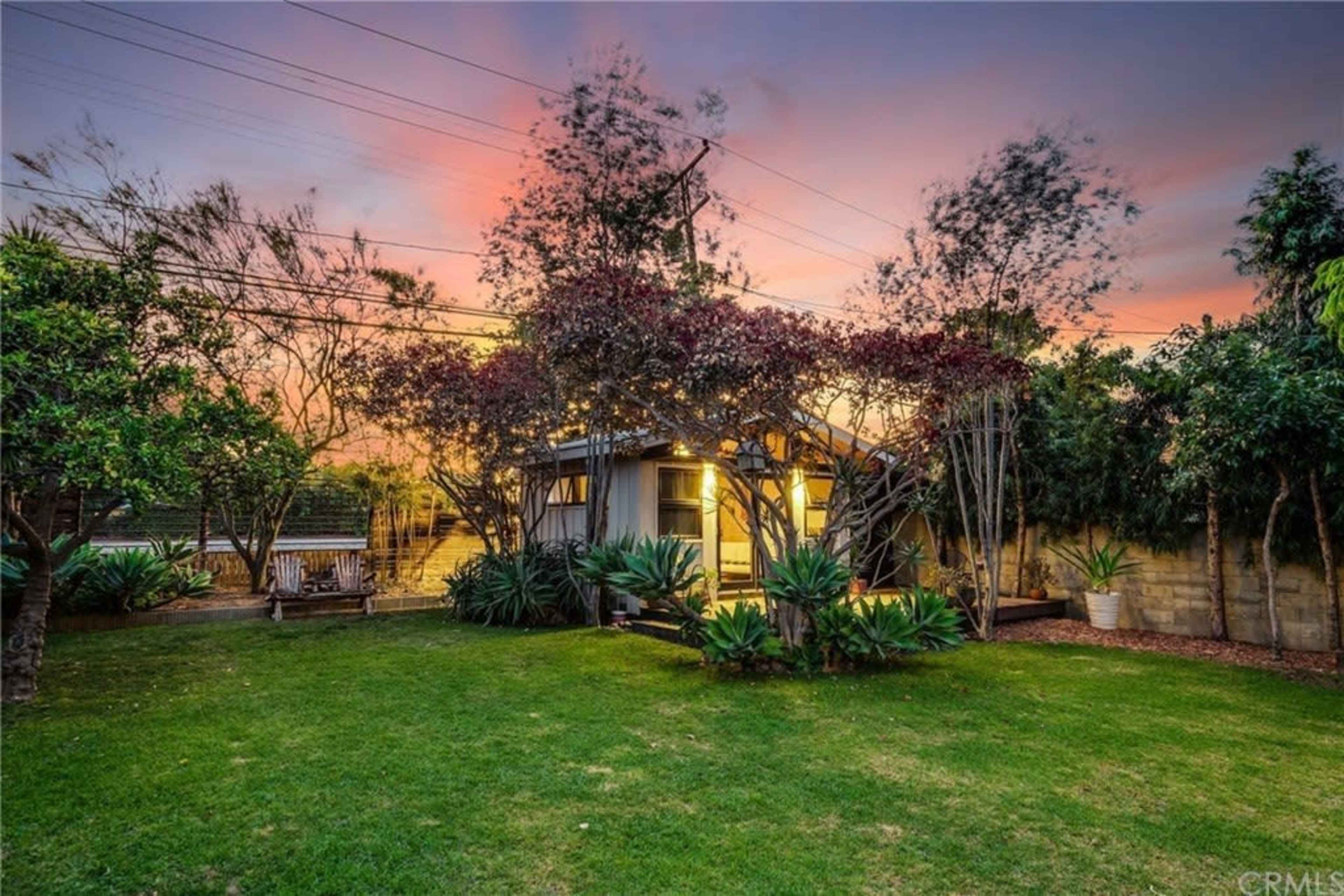 A small house with bright lights stands surrounded by greenery in a backyard at sunset.