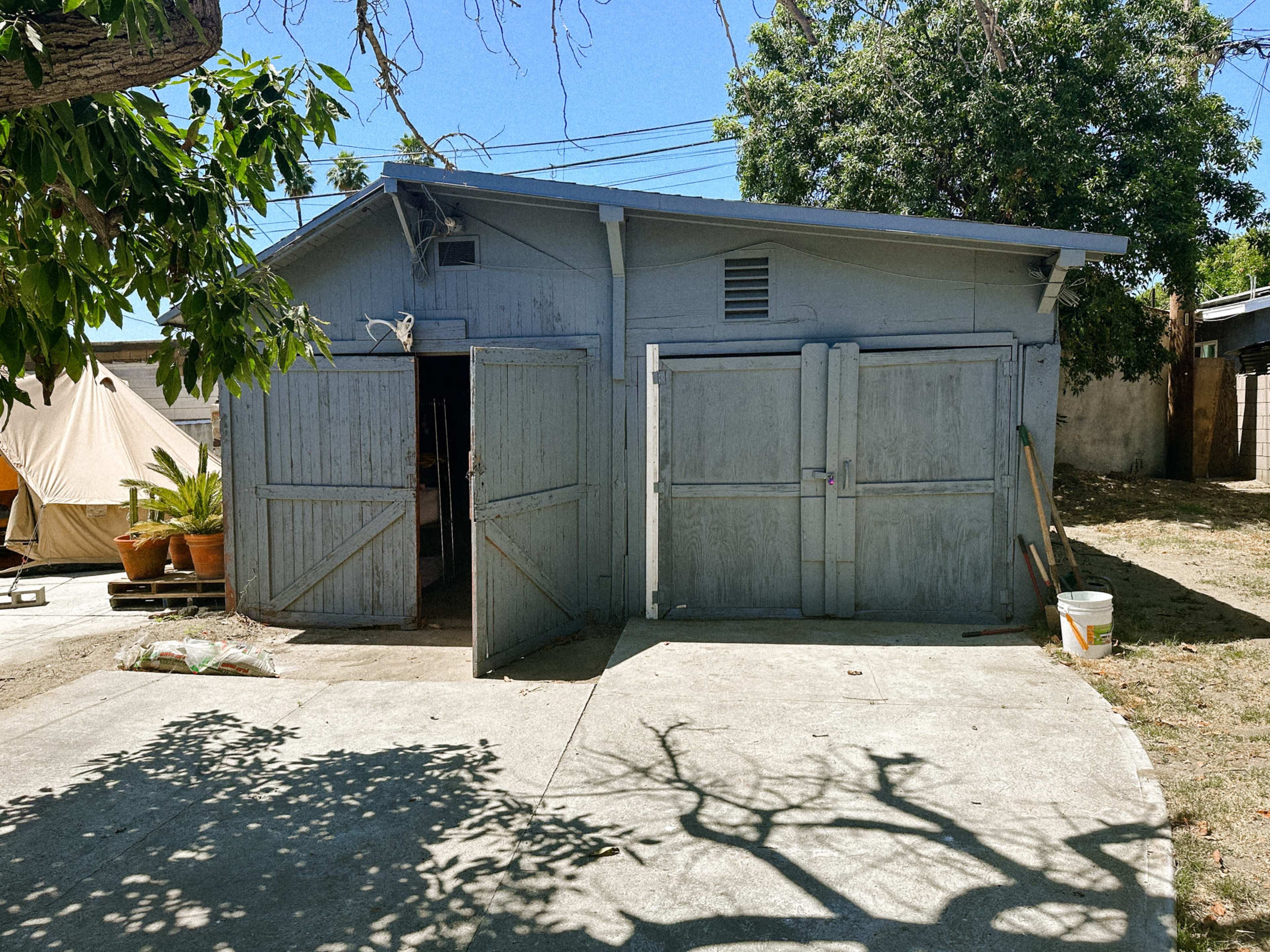 A weathered gray storage shed with double doors, set in a yard with a concrete surface and a few trees nearby.