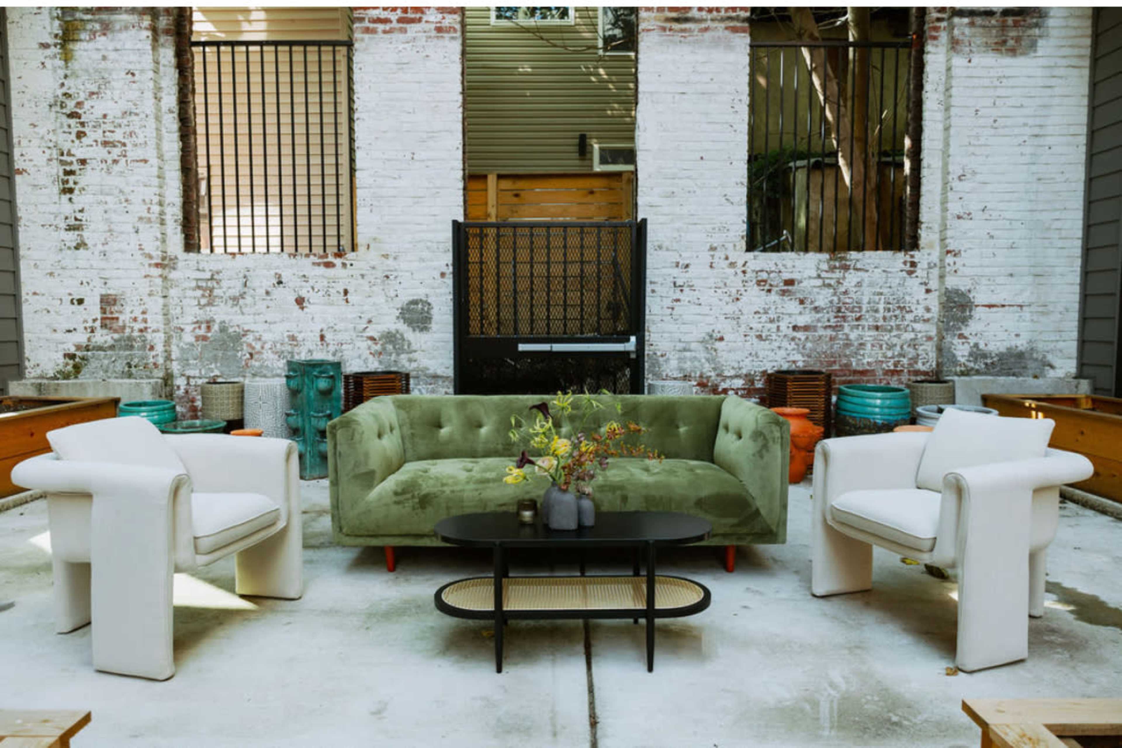 The scene features a green velvet sofa and two white armchairs arranged around a black coffee table in a rustic indoor space with exposed brick walls.