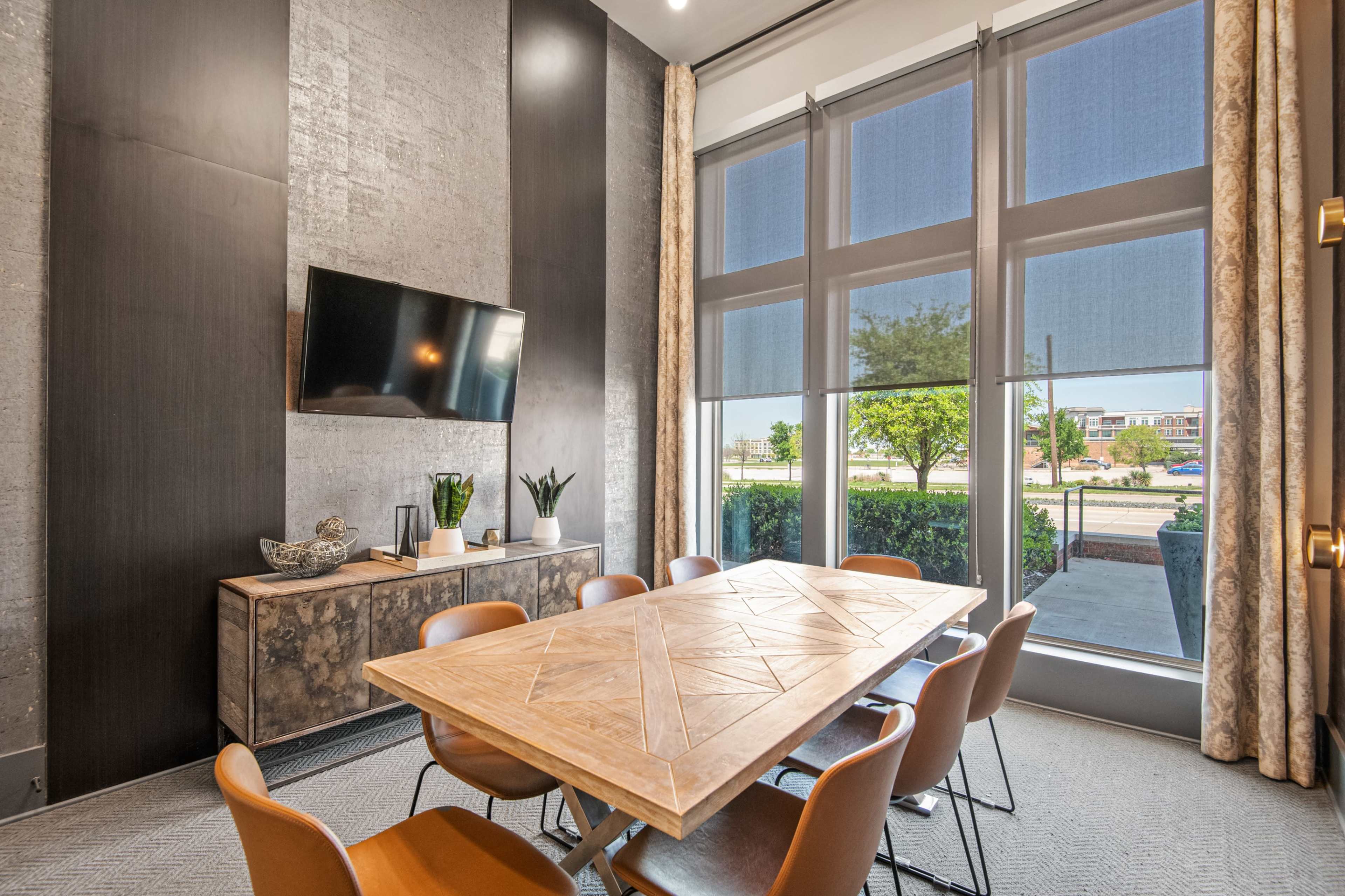 A modern meeting room features a large wooden table surrounded by orange chairs, a wall-mounted television, and large windows with decorative curtains.