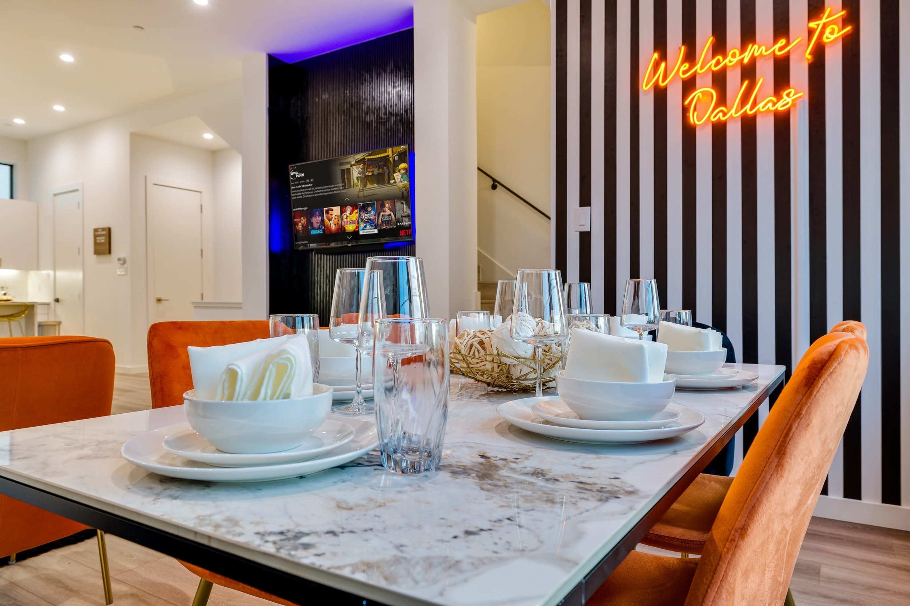 A dining table set with plates and glasses is positioned in a modern room featuring a neon sign that reads "Welcome to Dallas."