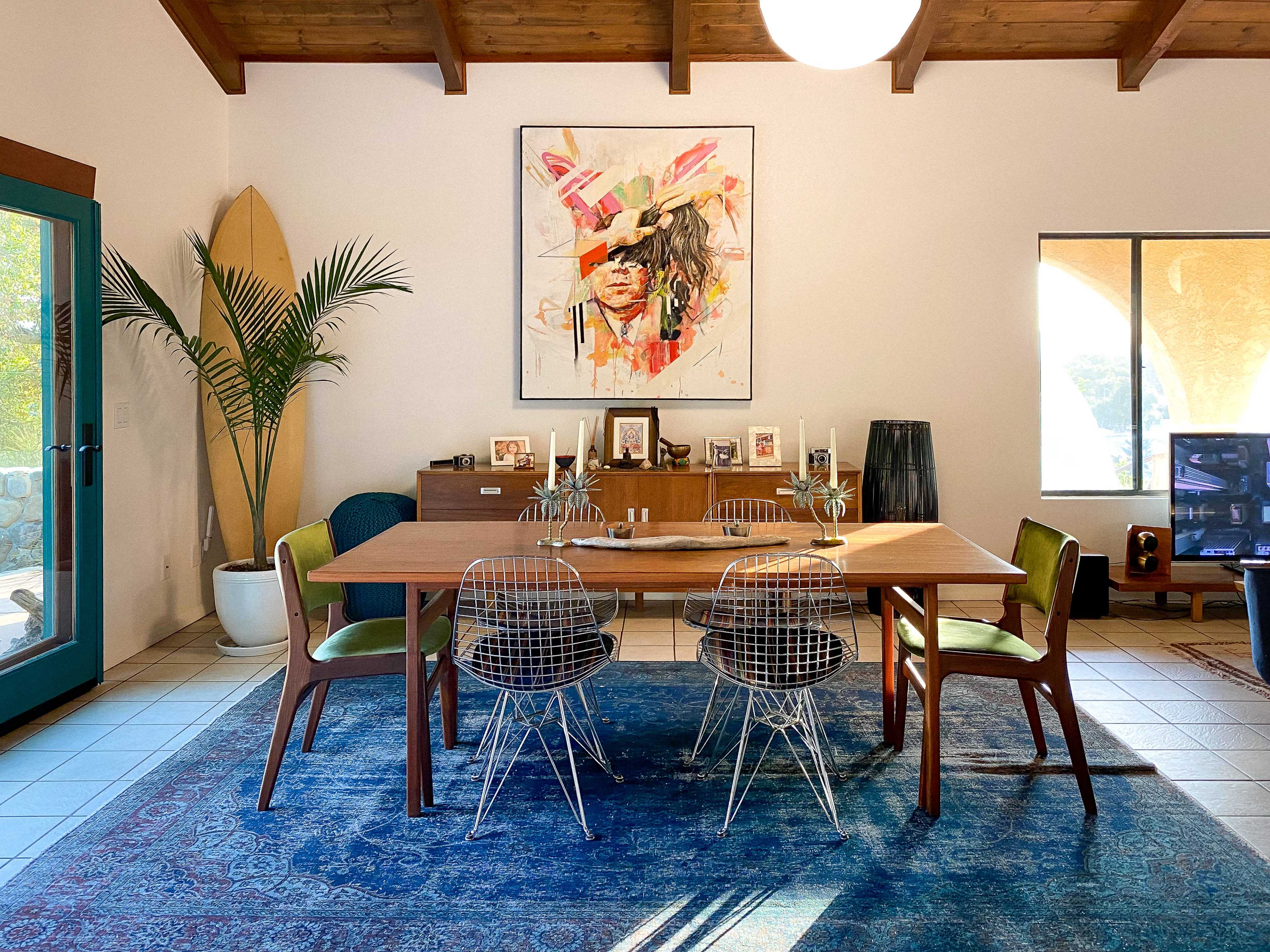 The image shows a modern dining space featuring a wooden table surrounded by wire chairs, a large artwork on the wall, and decorative elements including plants and a surfboard.