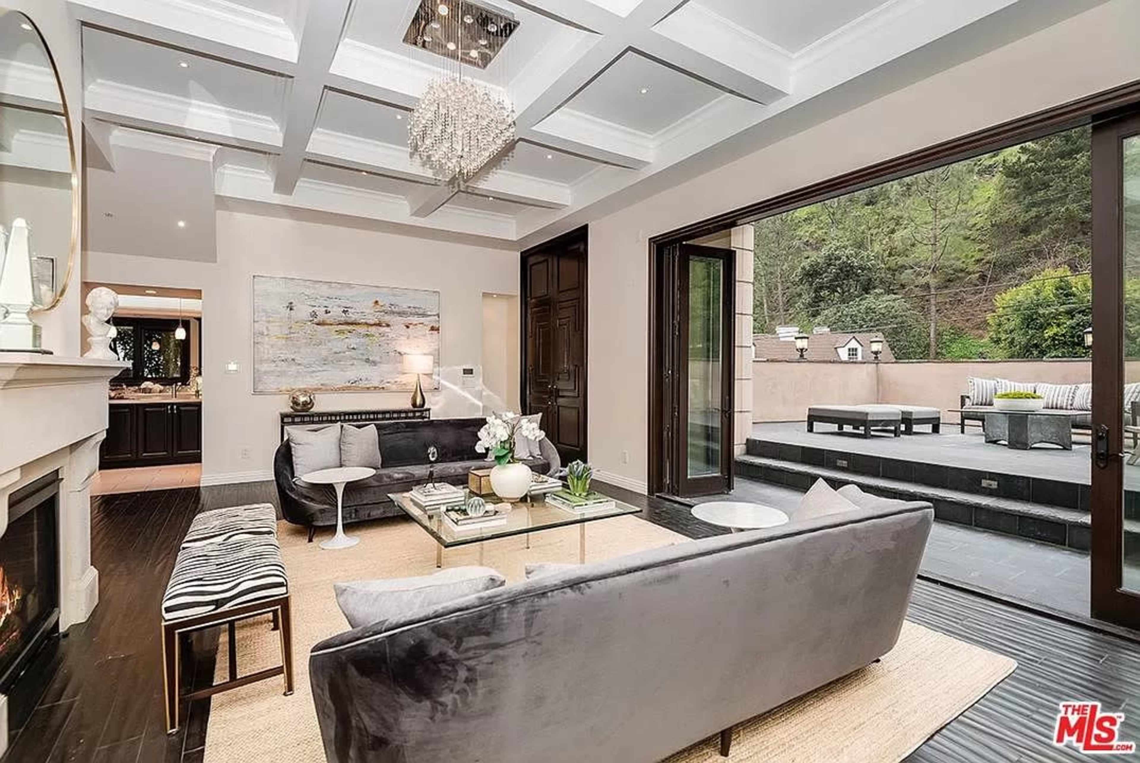 The living room features a gray sofa, a glass coffee table, and large sliding doors that open to an outdoor patio with a garden view.