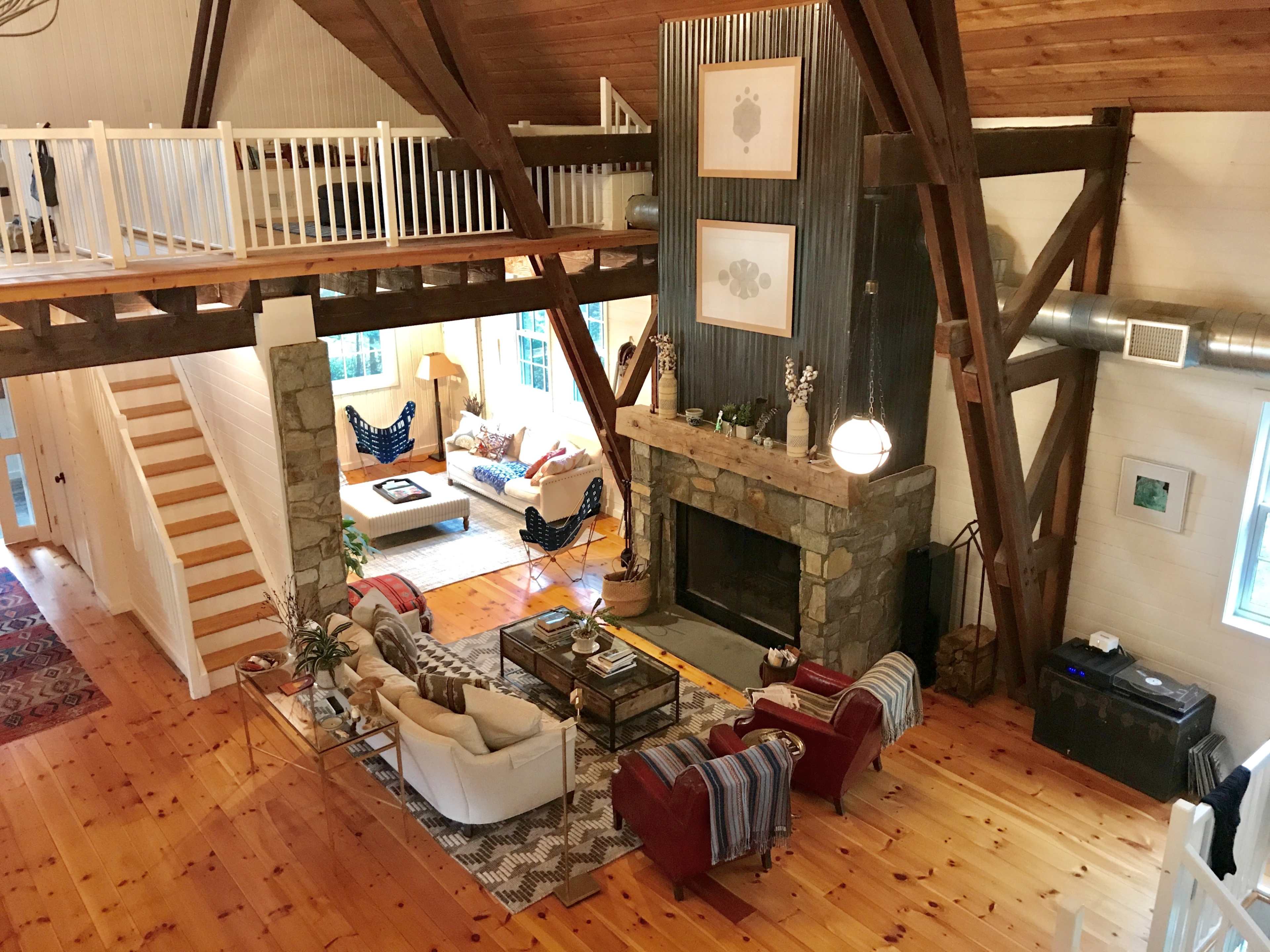 A spacious open-concept living area with a wooden interior, featuring a stone fireplace, a cozy seating arrangement, and a lofted section with a view of a staircase.