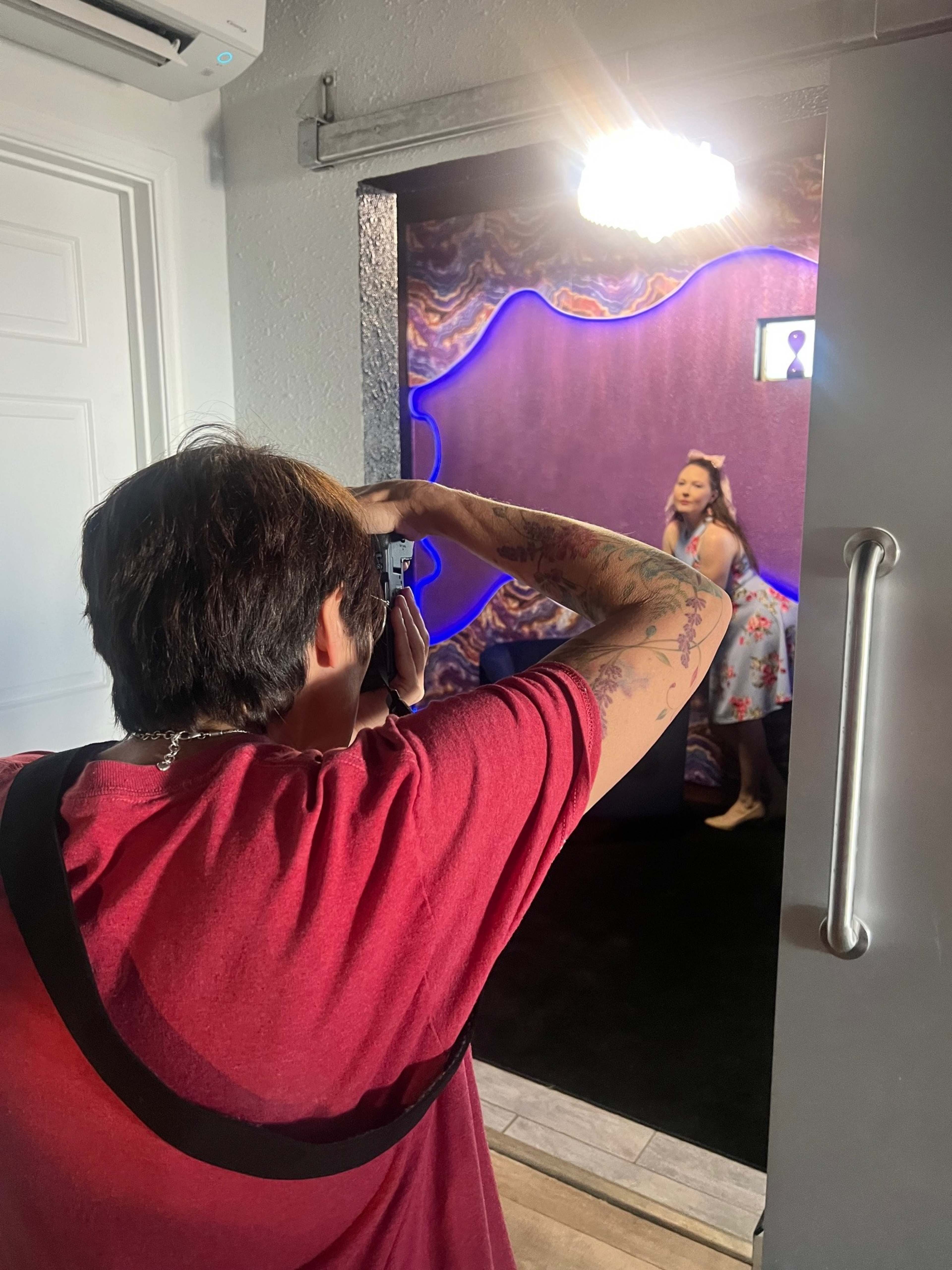 A person with a camera is photographing a woman posing in front of a colorful backdrop featuring purple and blue lighting.