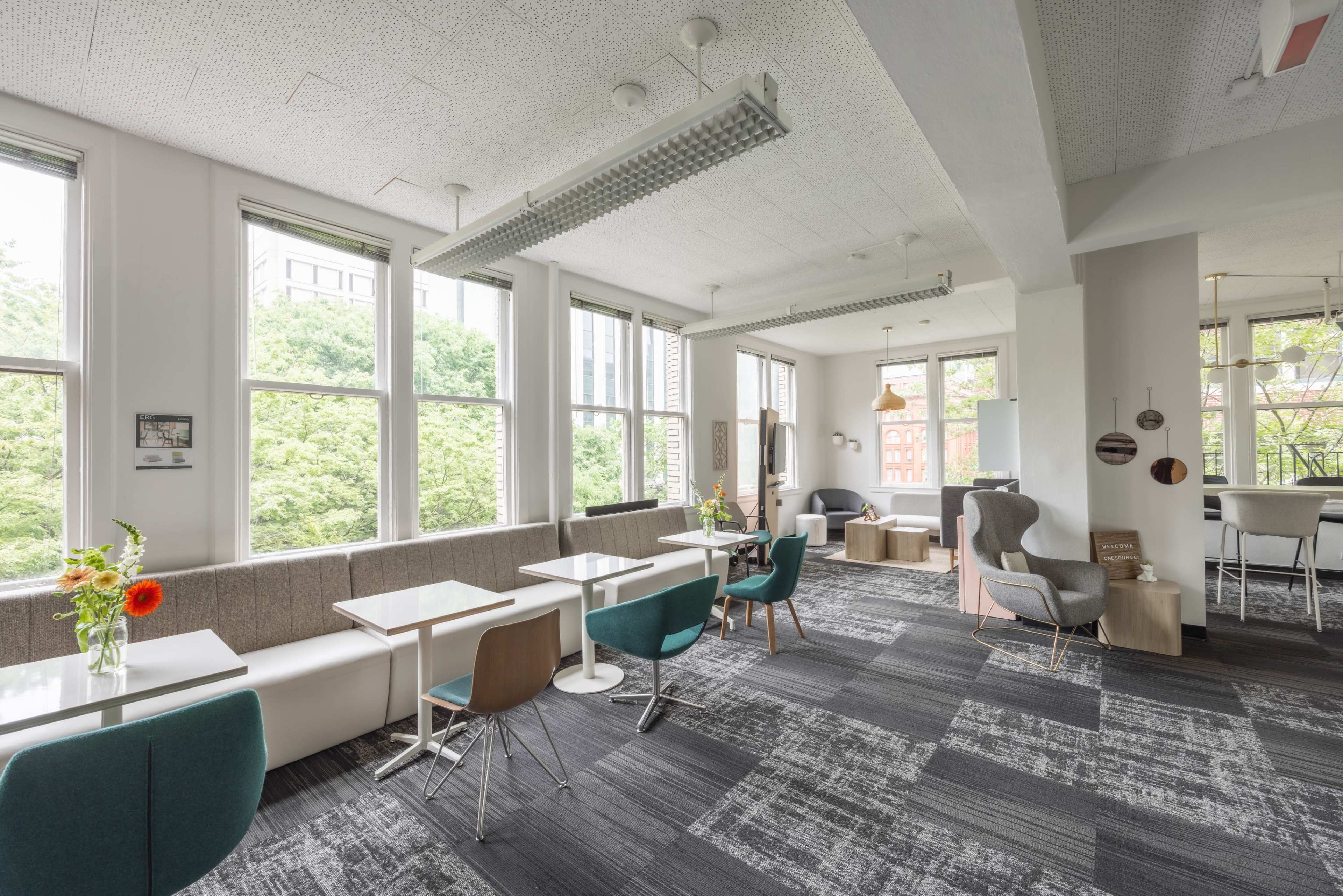 A modern workspace with several tables and chairs near large windows, featuring a mix of seating areas and greenery outside.
