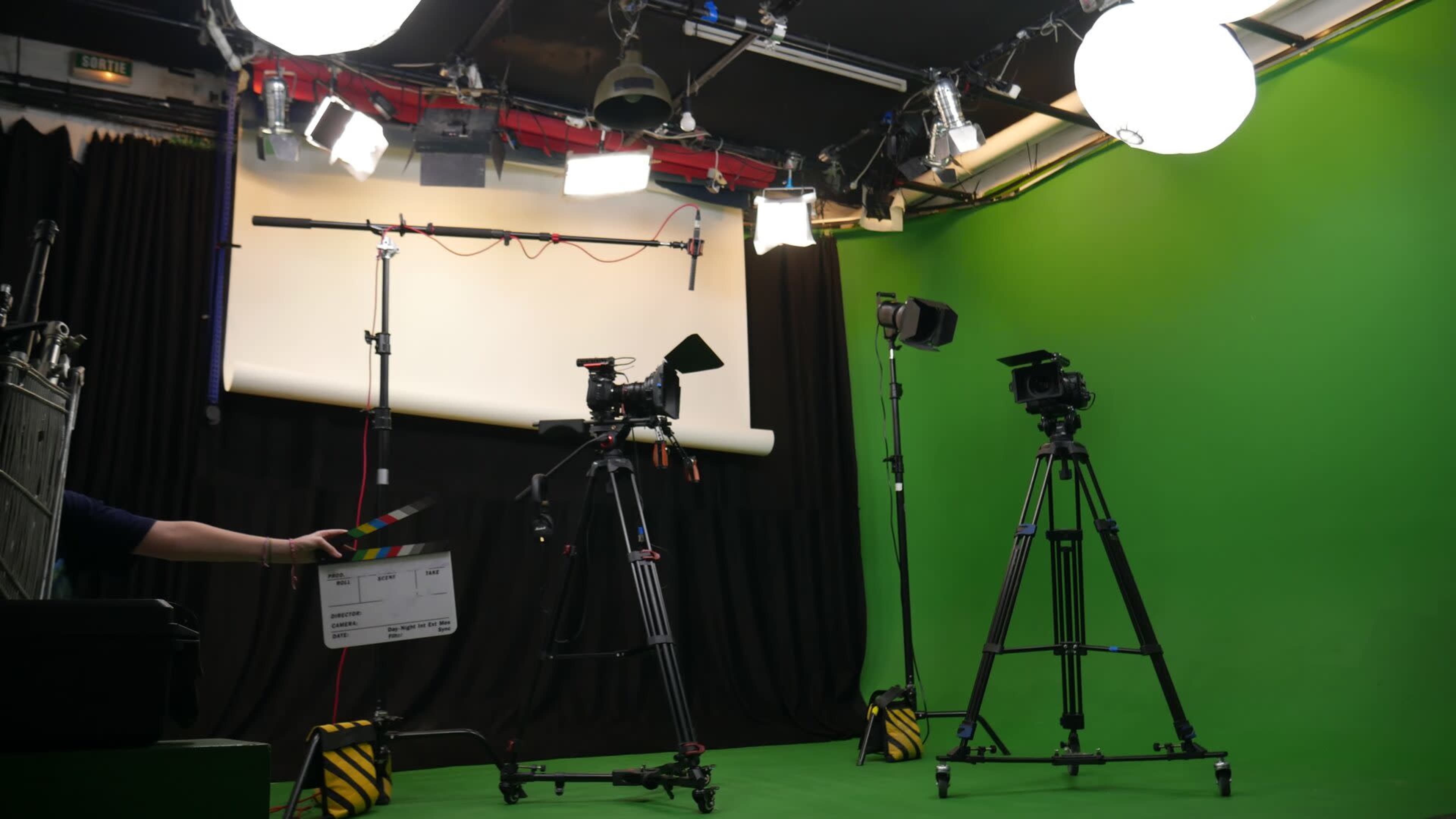 The image shows a film studio setup with two tripods holding cameras, a clapperboard, and lighting equipment against a green chroma key backdrop.