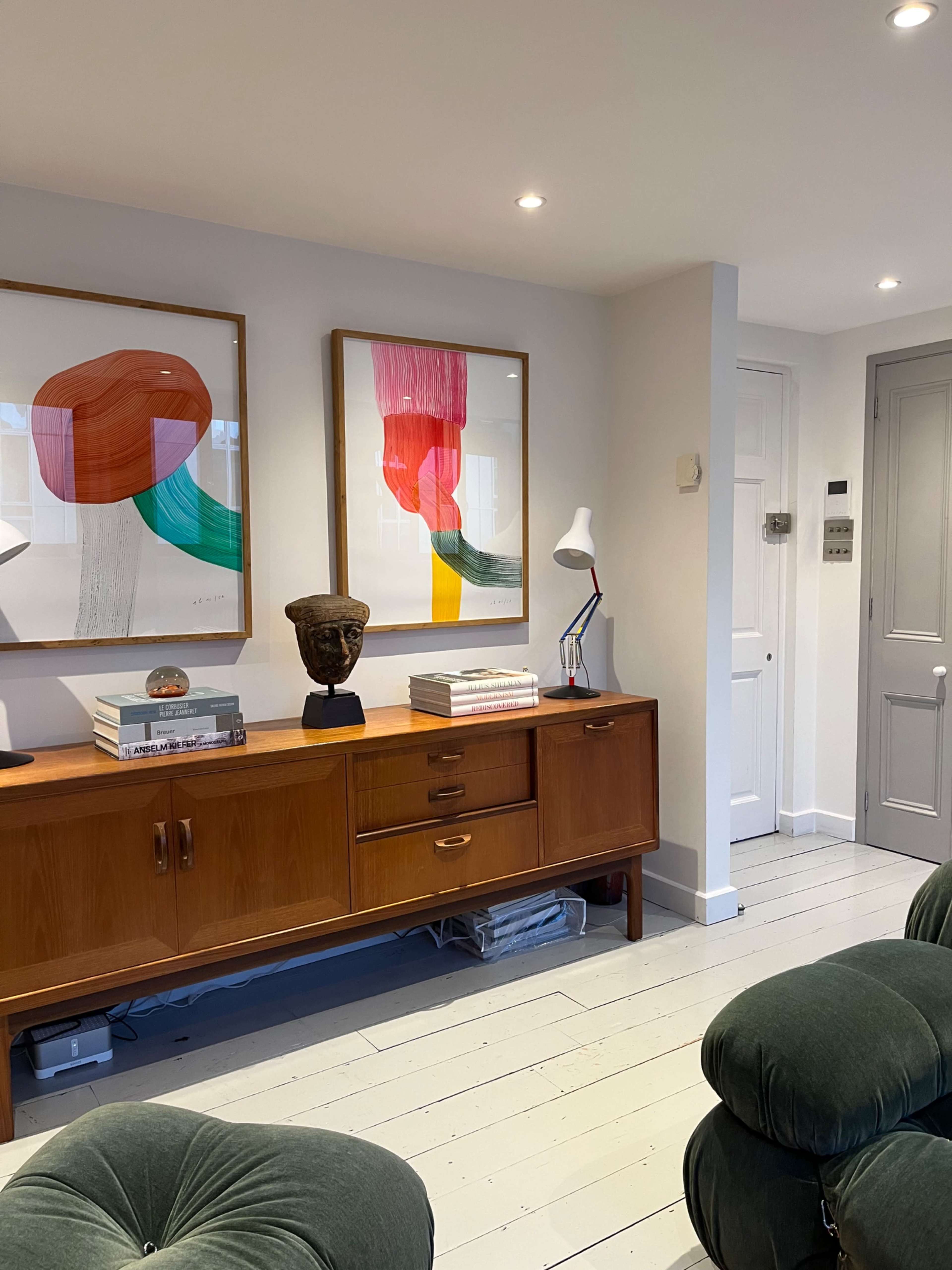 The image shows a modern living space featuring a wooden sideboard, two colorful framed artworks on the wall, a sculpture on the sideboard, and a green armchair.