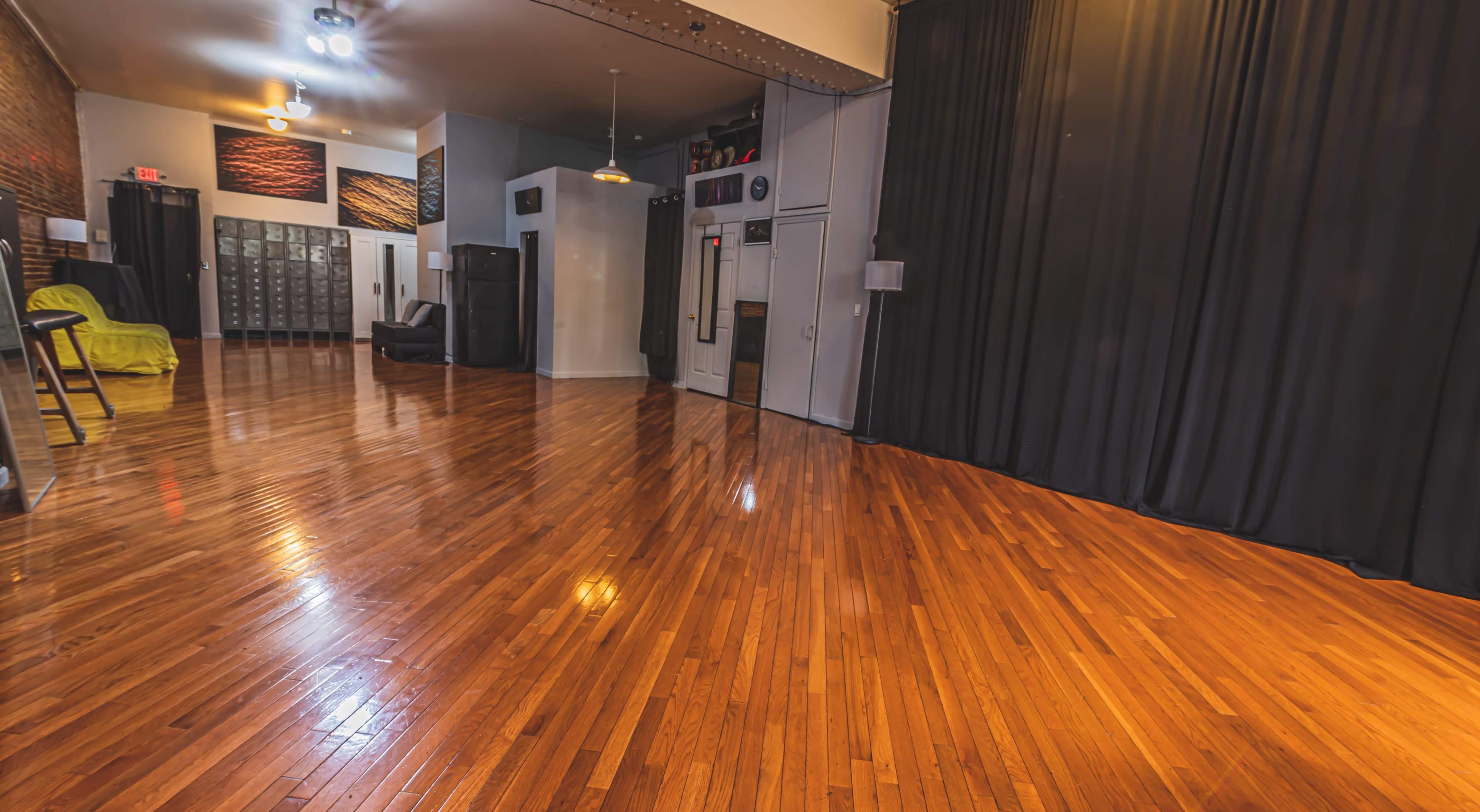 The image shows a spacious room with polished wooden floors, black curtains on one side, and various furniture and lockers along the walls.