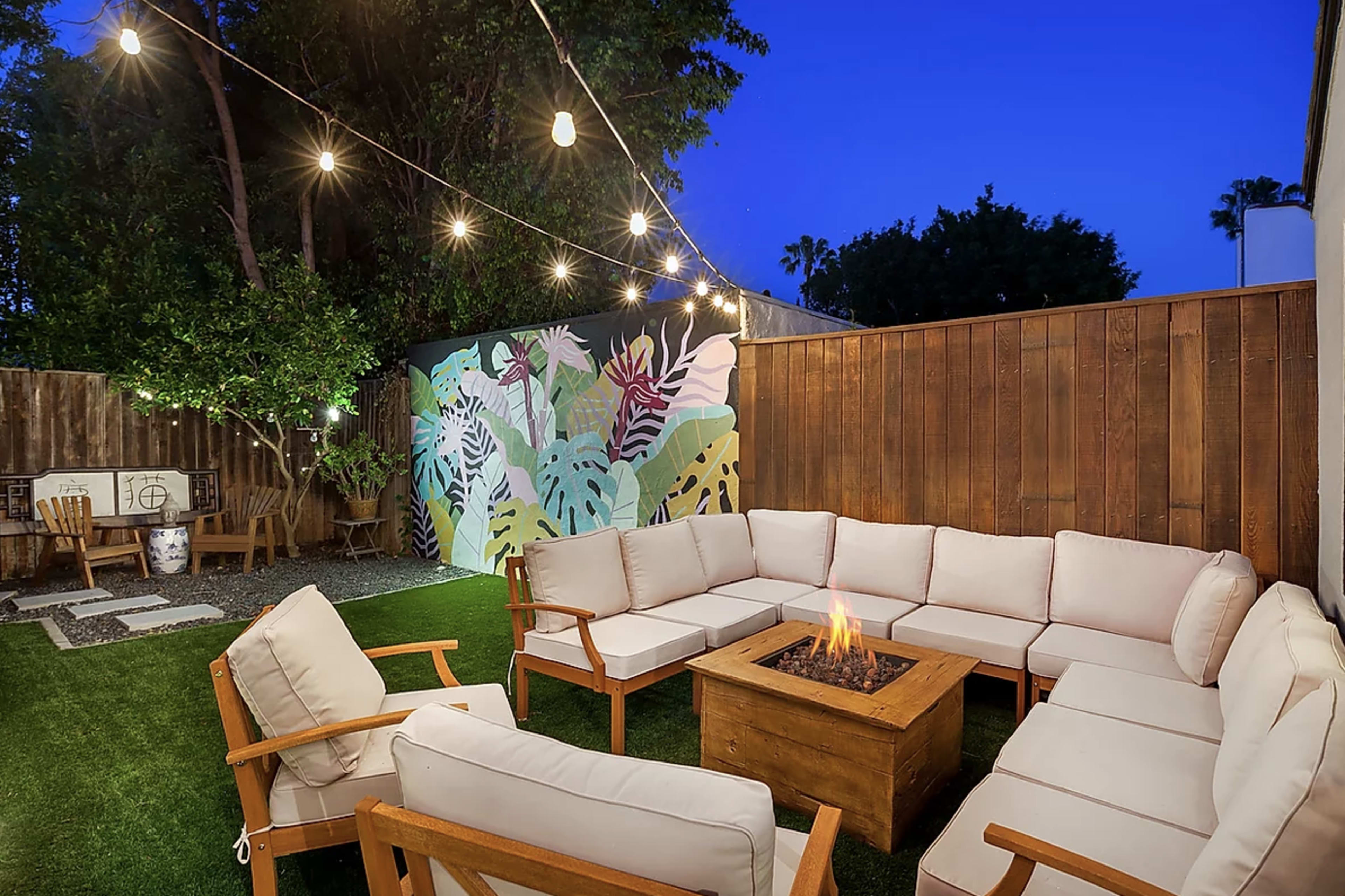 The image shows an outdoor seating area with a fire pit surrounded by a circular arrangement of cushioned chairs and a colorful mural on the wall.