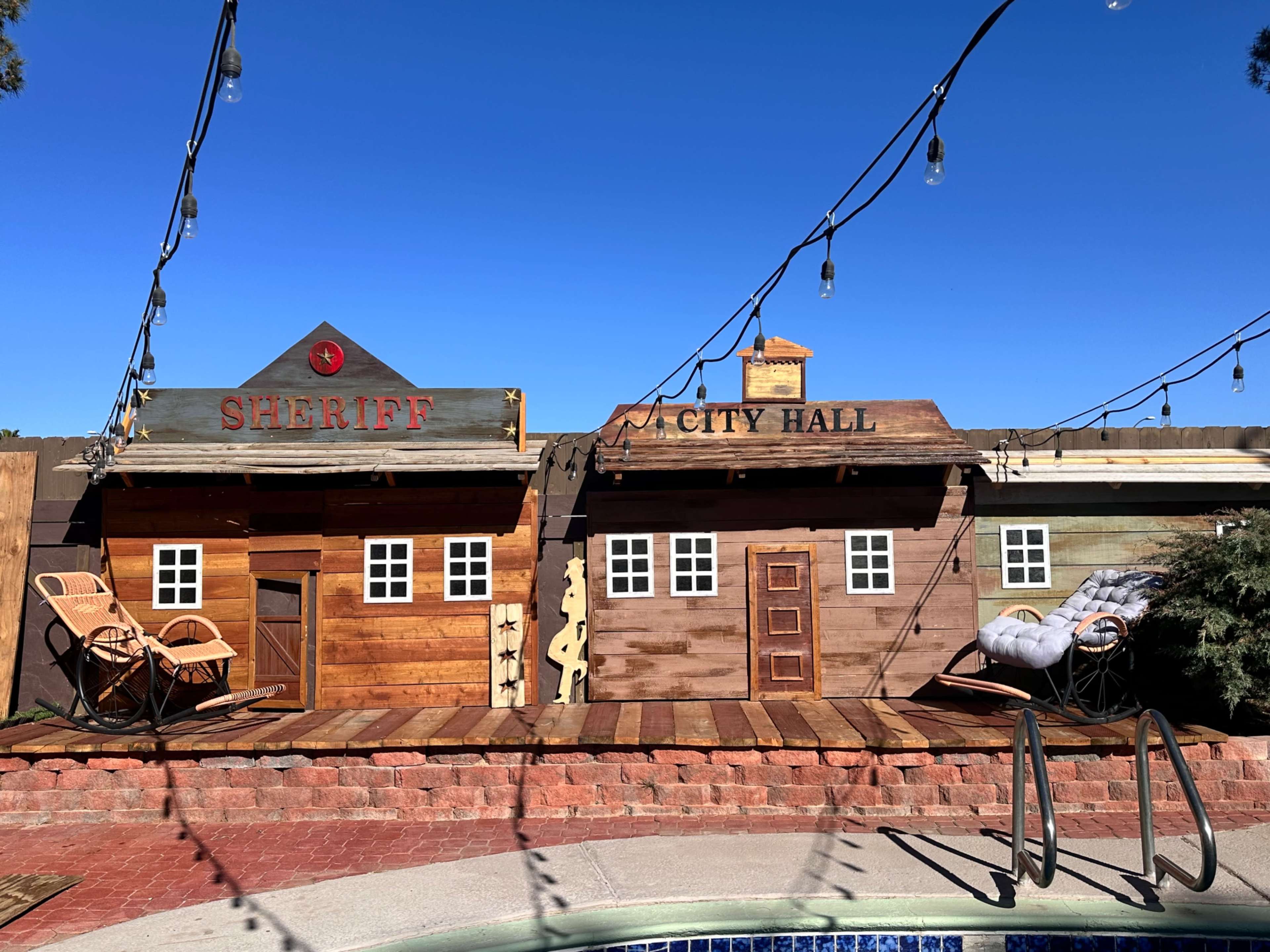 The scene shows two wooden buildings labeled "SHERIFF" and "CITY HALL" positioned near a pool, with string lights overhead and lounge chairs in front.