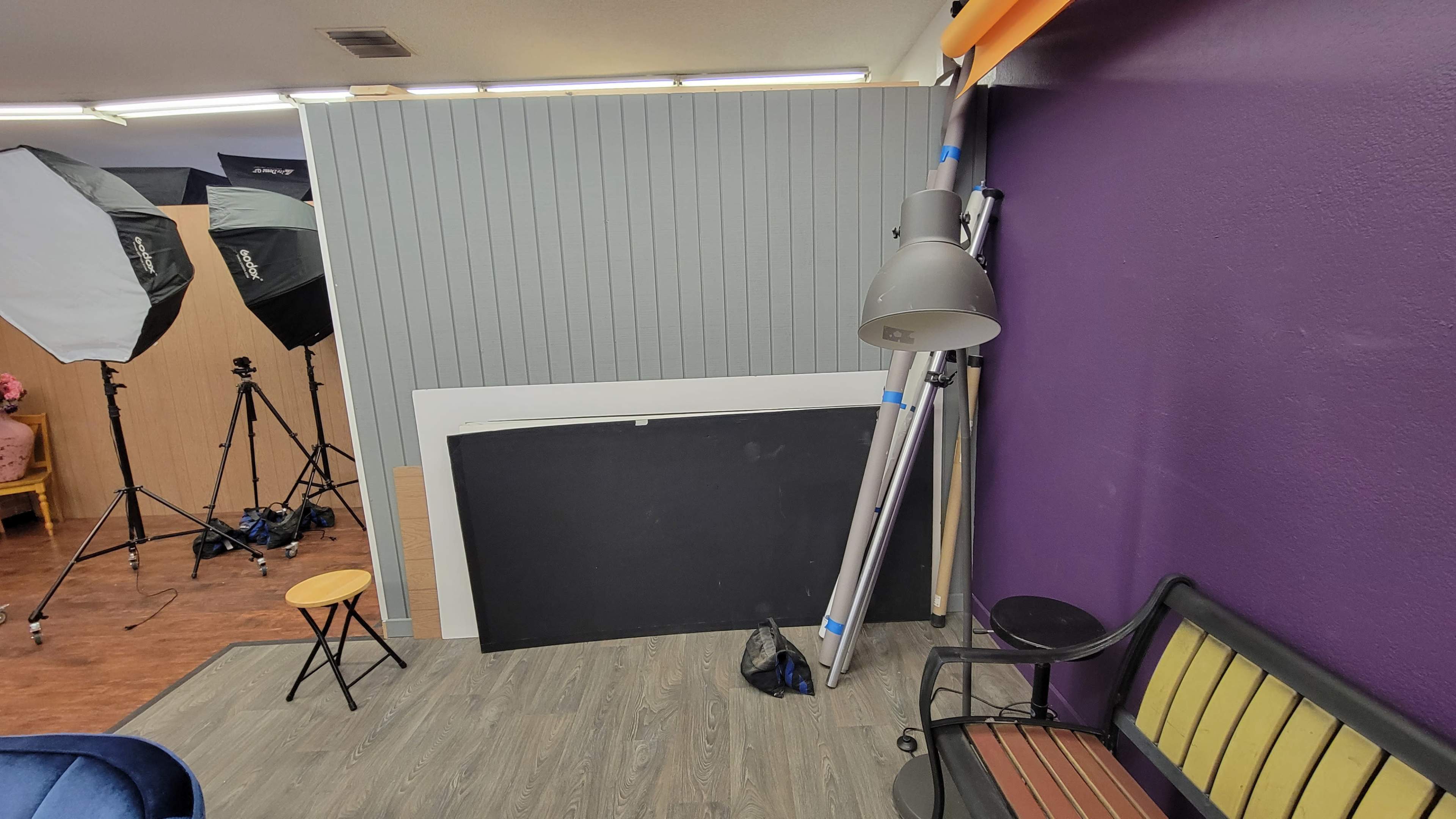 A room setup with a purple wall, a gray panel, photography equipment including softboxes, a tripod, and a floor lamp beside a seating area.