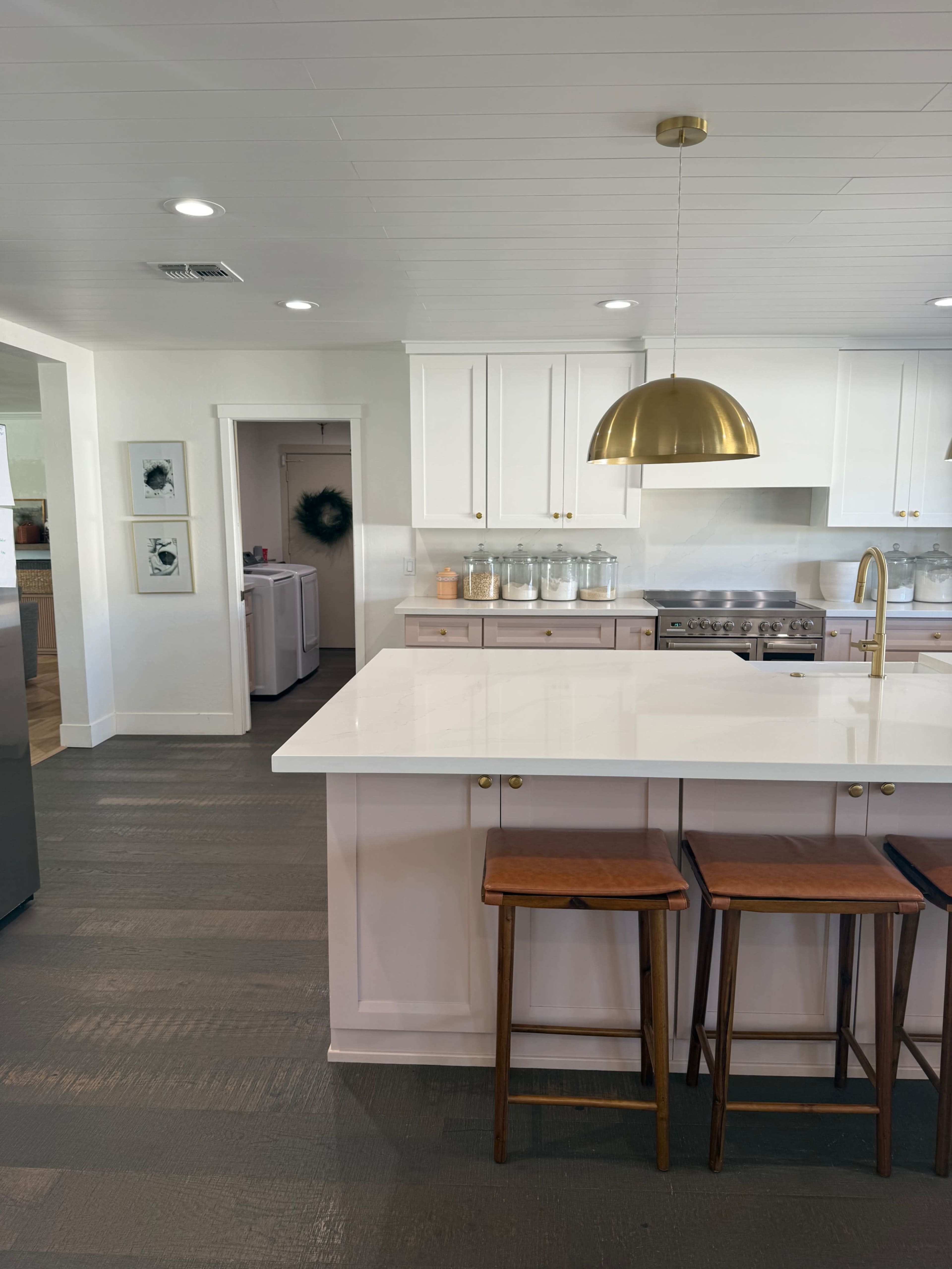 The image shows a modern kitchen with white cabinetry, a large island featuring a white countertop, two wooden stools, and a gold pendant light.
