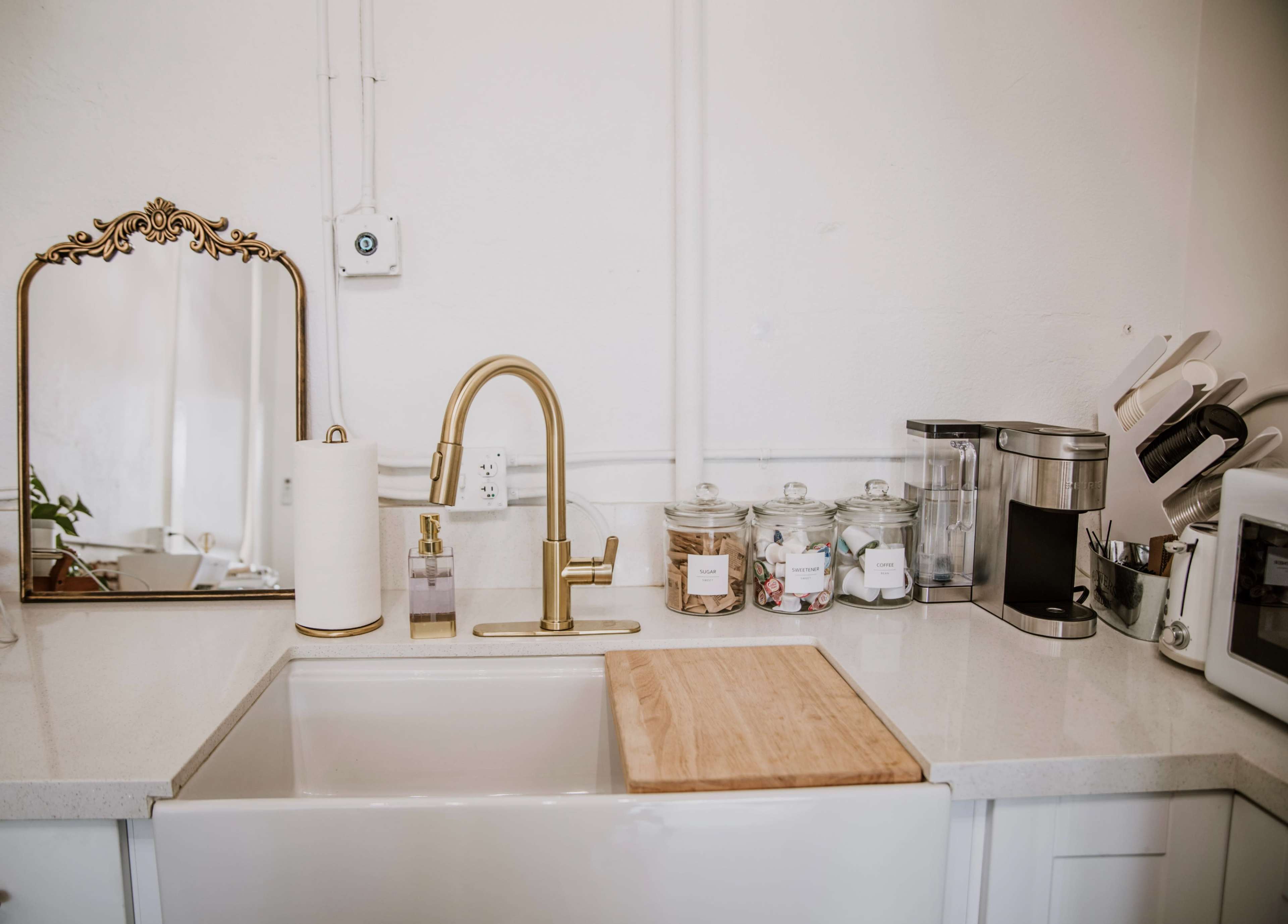 A kitchen countertop features a sink, gold faucet, mirror, and various kitchen appliances and jars.