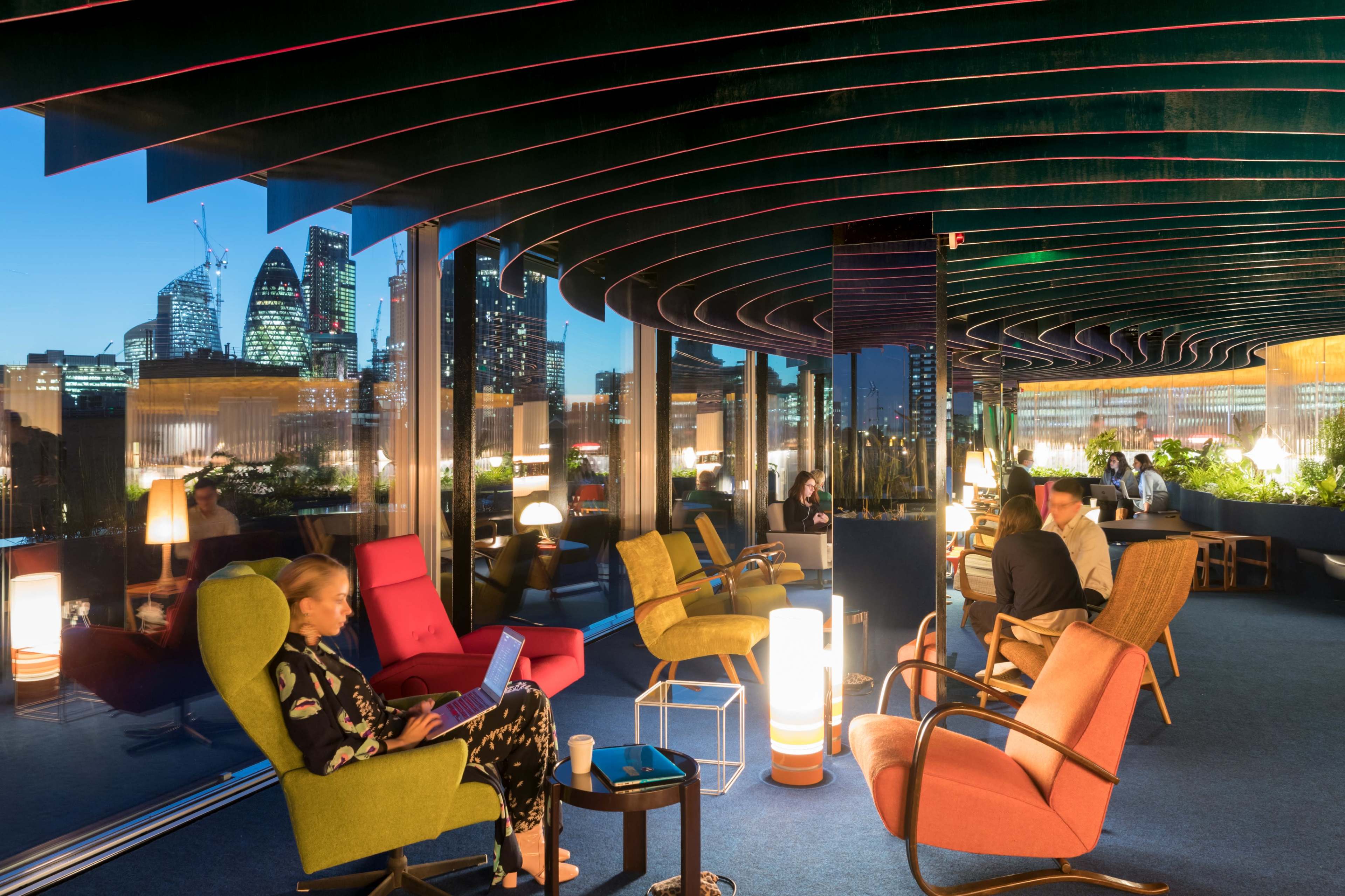 A modern lounge features colorful seating arrangements with large windows overlooking a city skyline at dusk.