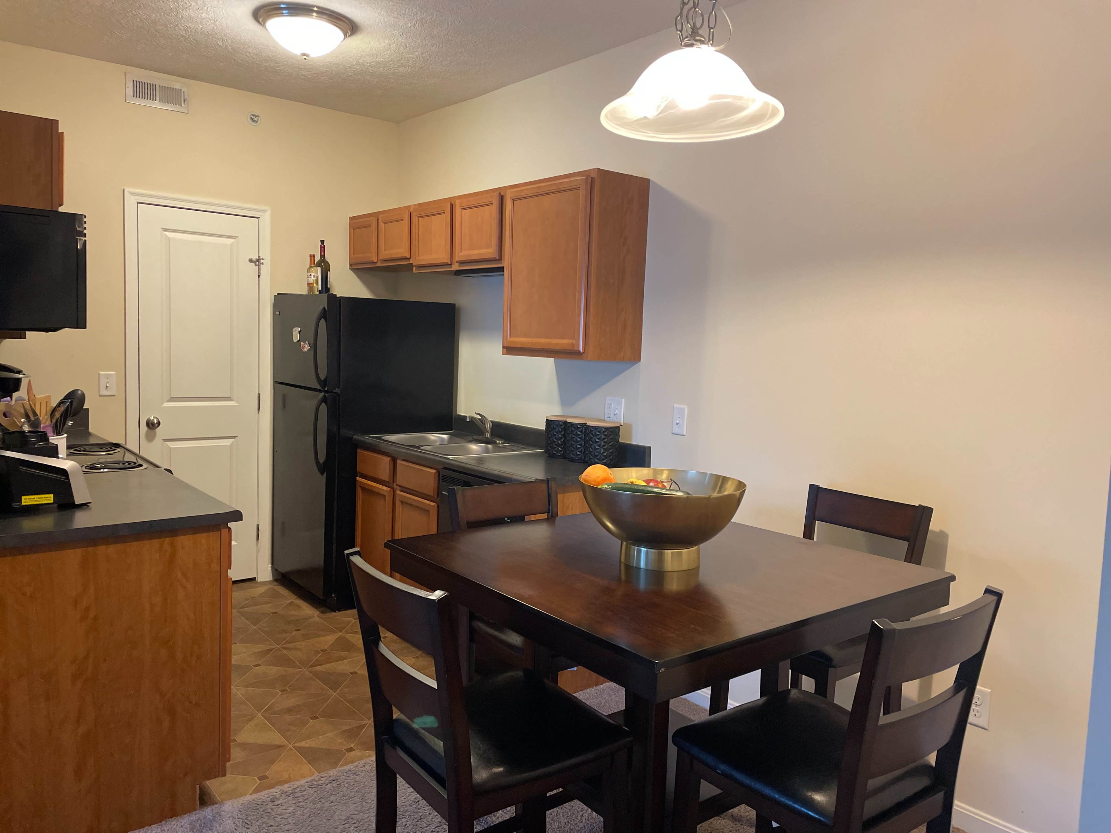 A kitchen and dining area featuring wooden cabinets, a black refrigerator, and a table with chairs, illuminated by a hanging light fixture.