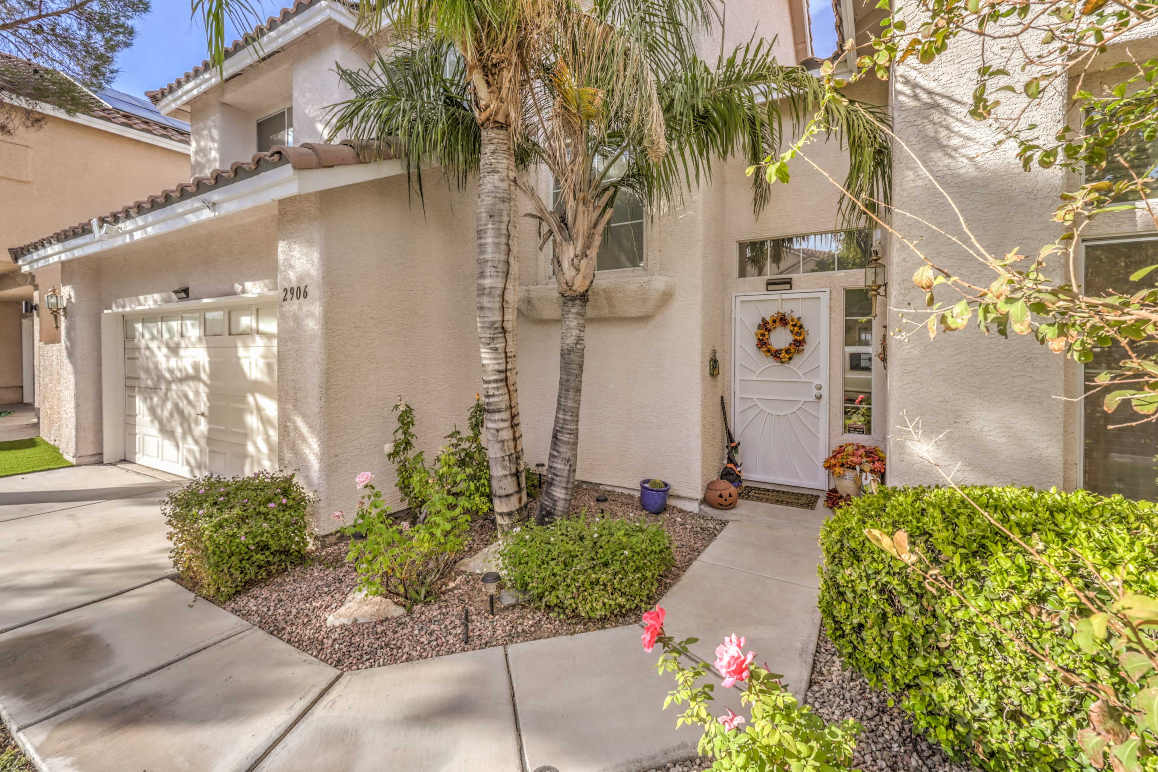 The entrance of a house with a decorative door, surrounded by palm trees and greenery, along with a pathway and flower beds.